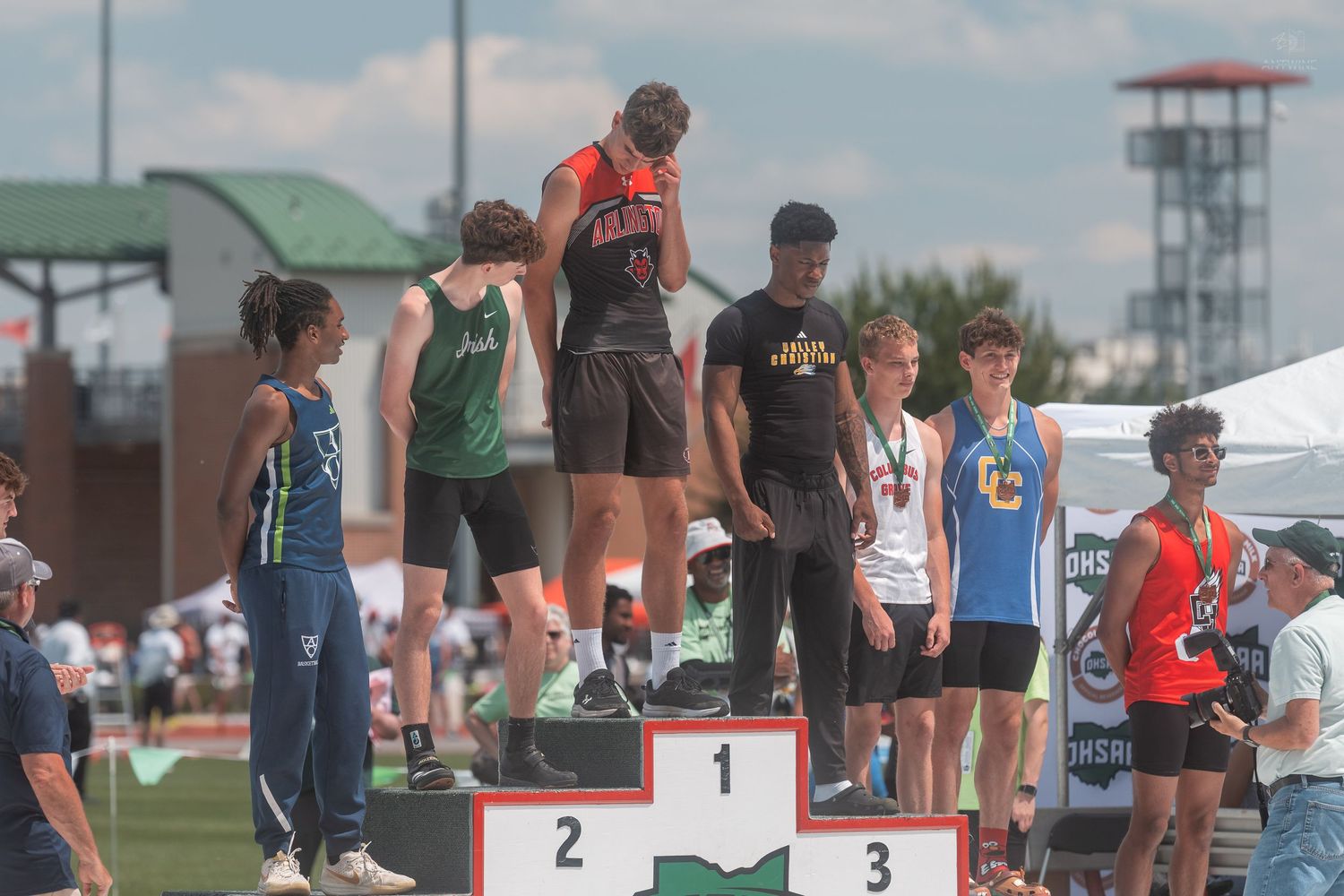 Athletes stand on a podium at a track and field competition, with the first-place winner elevated on the center platform.