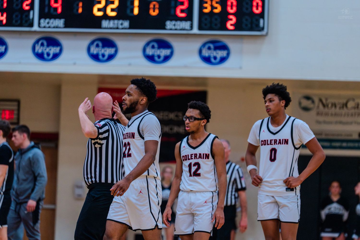 Colerain high school basketball players huddle on court during a game with scoreboard visible in background.