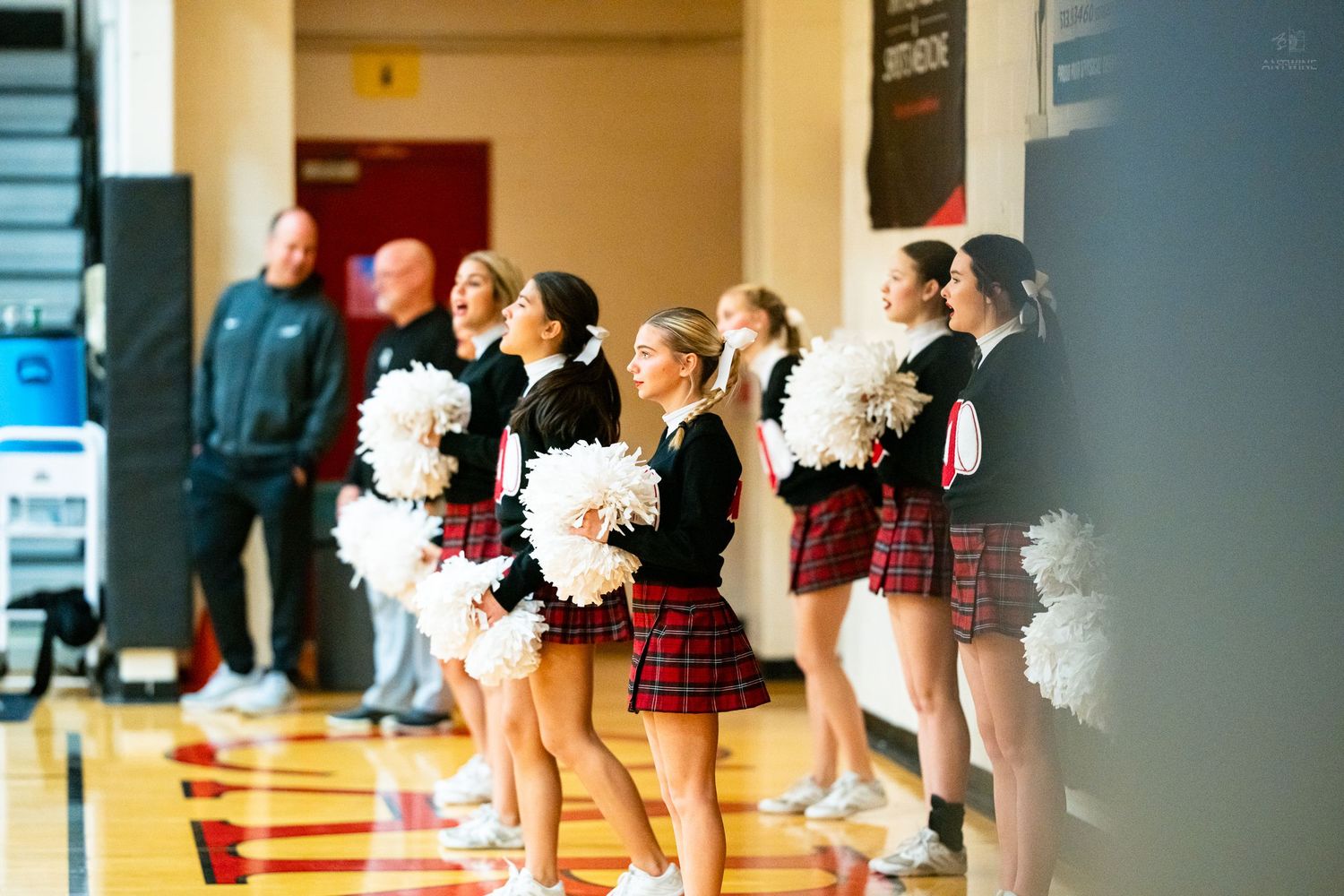 High school cheerleaders in plaid skirts hold white pom-poms while cheering at a basketball game.
