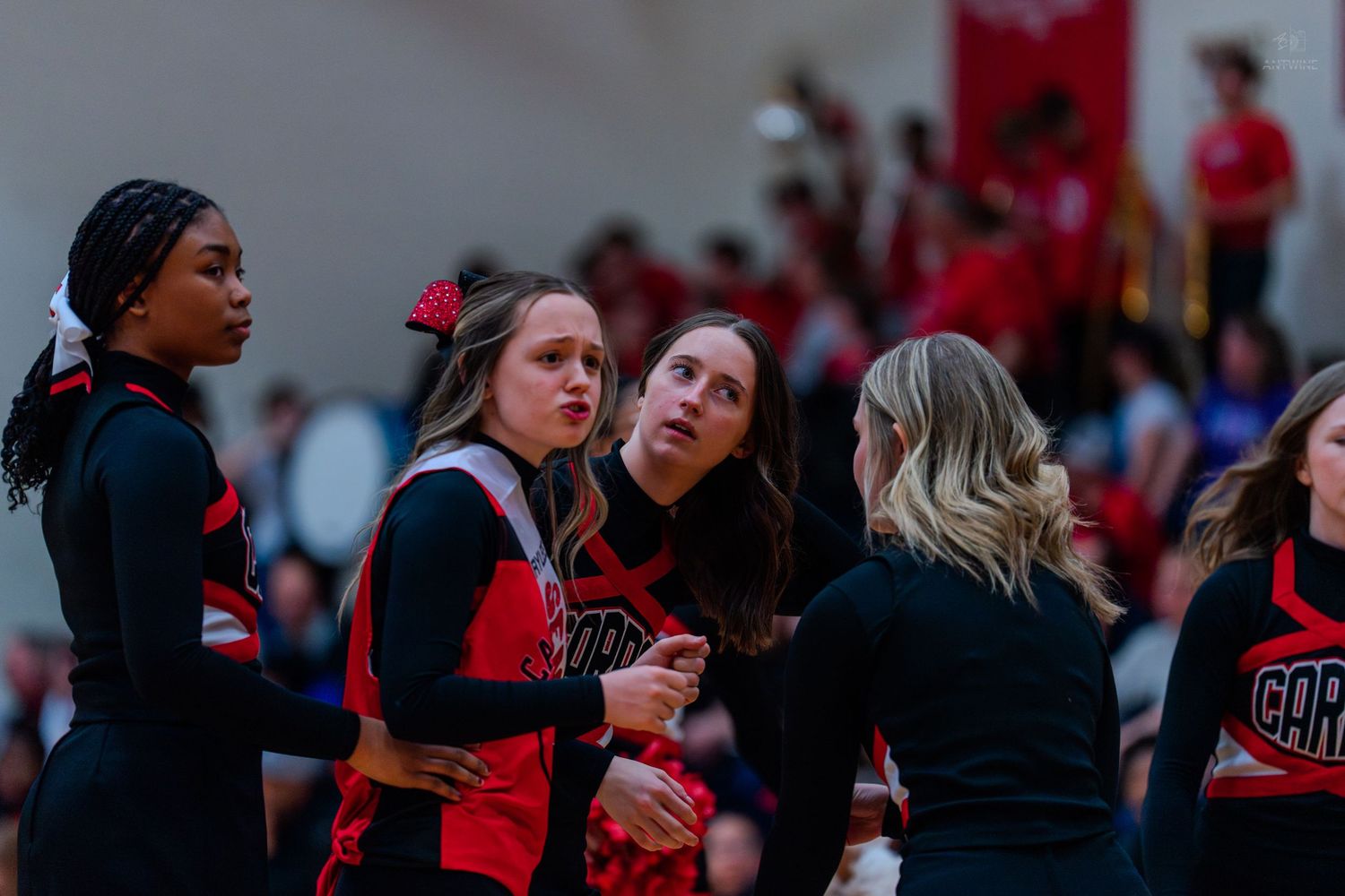 High school cheerleaders in red and black uniforms receive coaching during a basketball game timeout.