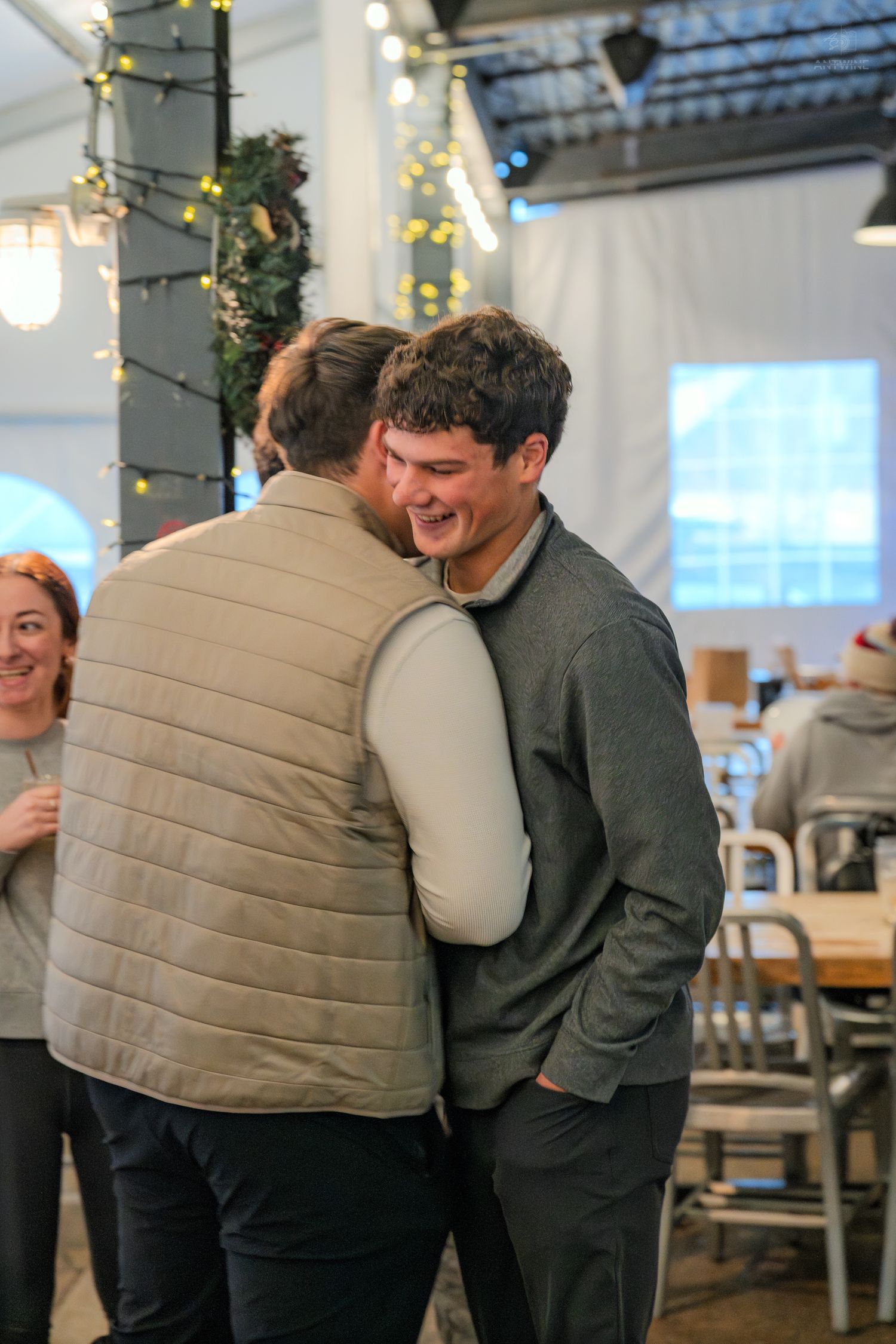 Two men share a warm embrace and laugh together at a festive indoor event with string lights in the background.