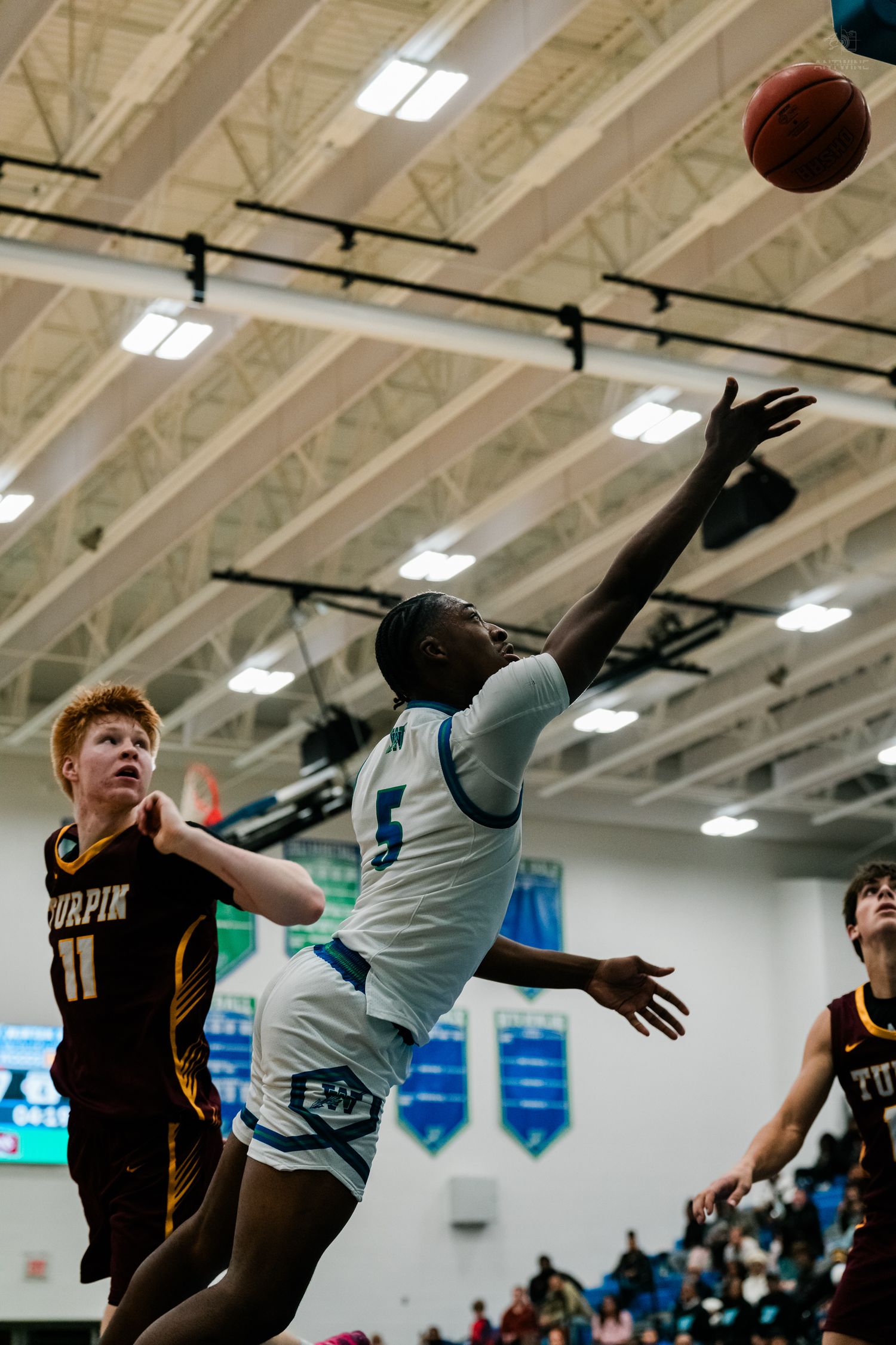 Player number 5 in white extends for a difficult layup attempt against Turpin defenders in a high school basketball game.