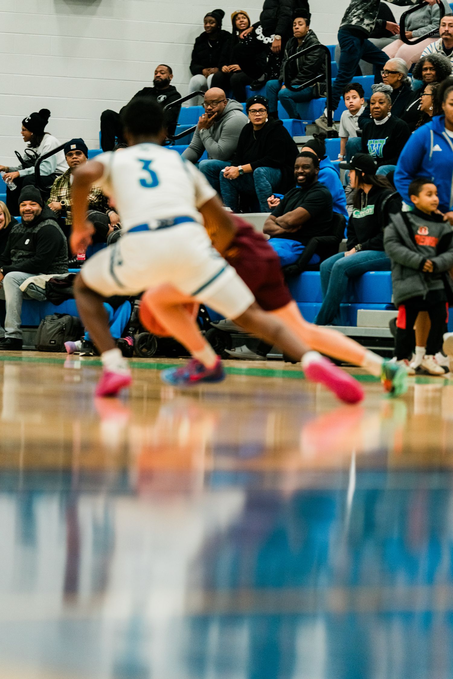 High school basketball player #3 in white uniform dribbles toward basket with crowd watching in bleachers.