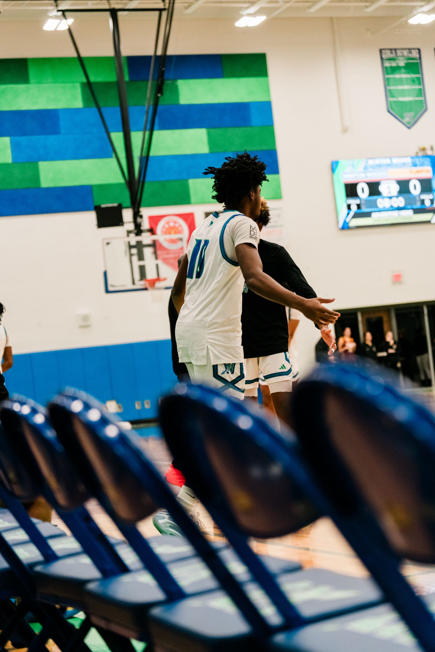High school basketball player #11 in white uniform walks near courtside chairs in colorful gymnasium.