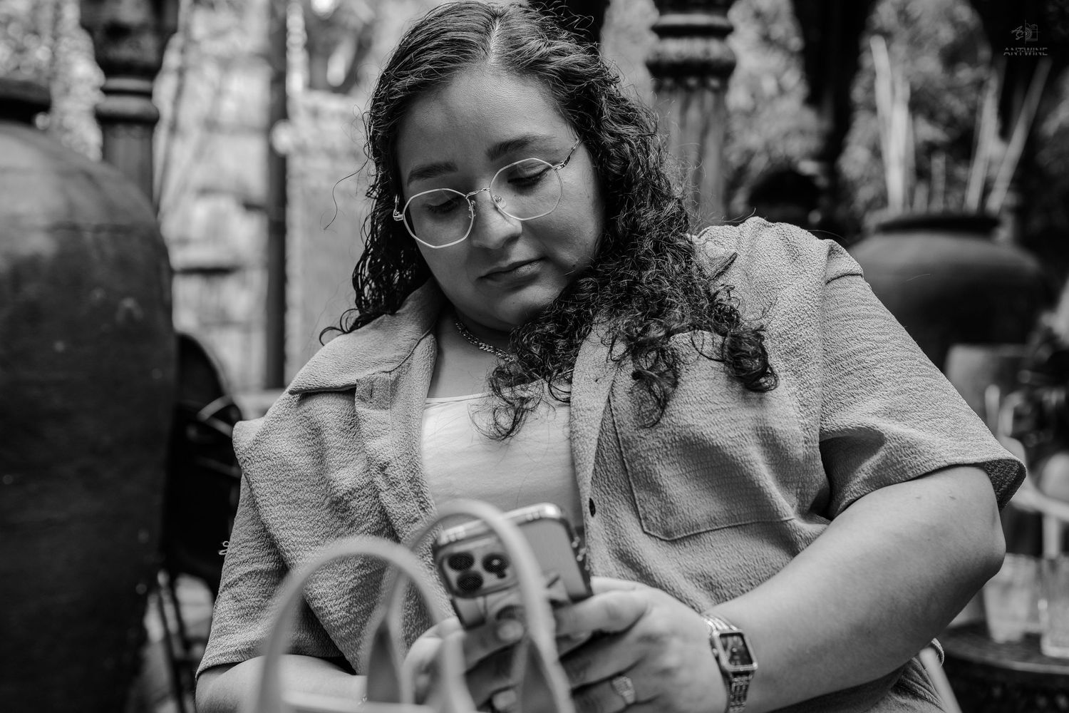Black and white portrait of curly-haired woman with glasses looking at phone outdoors.