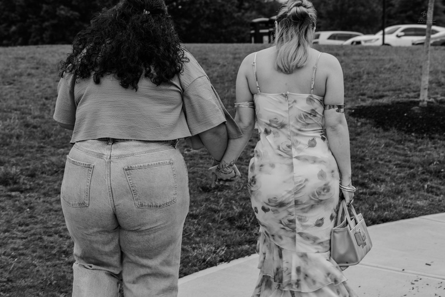 Black and white photo of two women holding hands while walking along a sidewalk in a park.