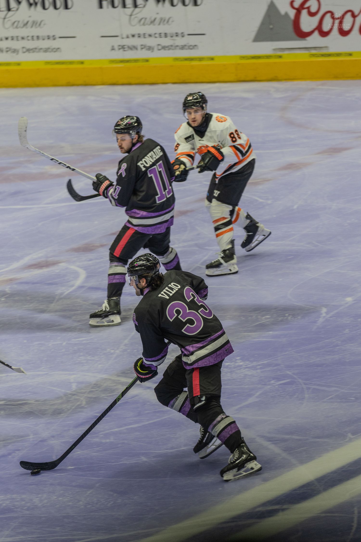 Two hockey players in purple jerseys compete against an orange-uniformed opponent during a minor league game.