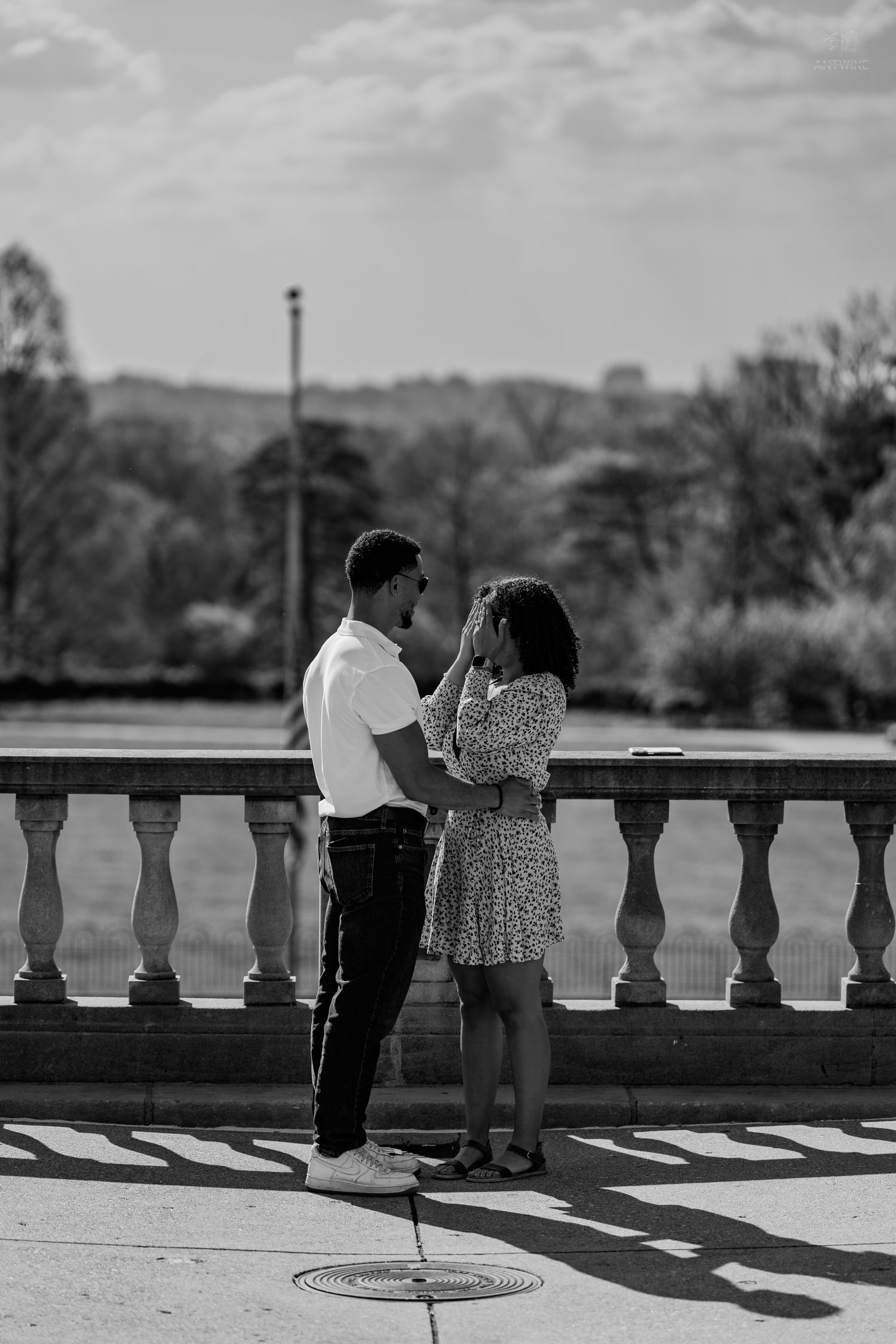 Black and white photo of couple holding hands by ornate stone railing in outdoor park setting.