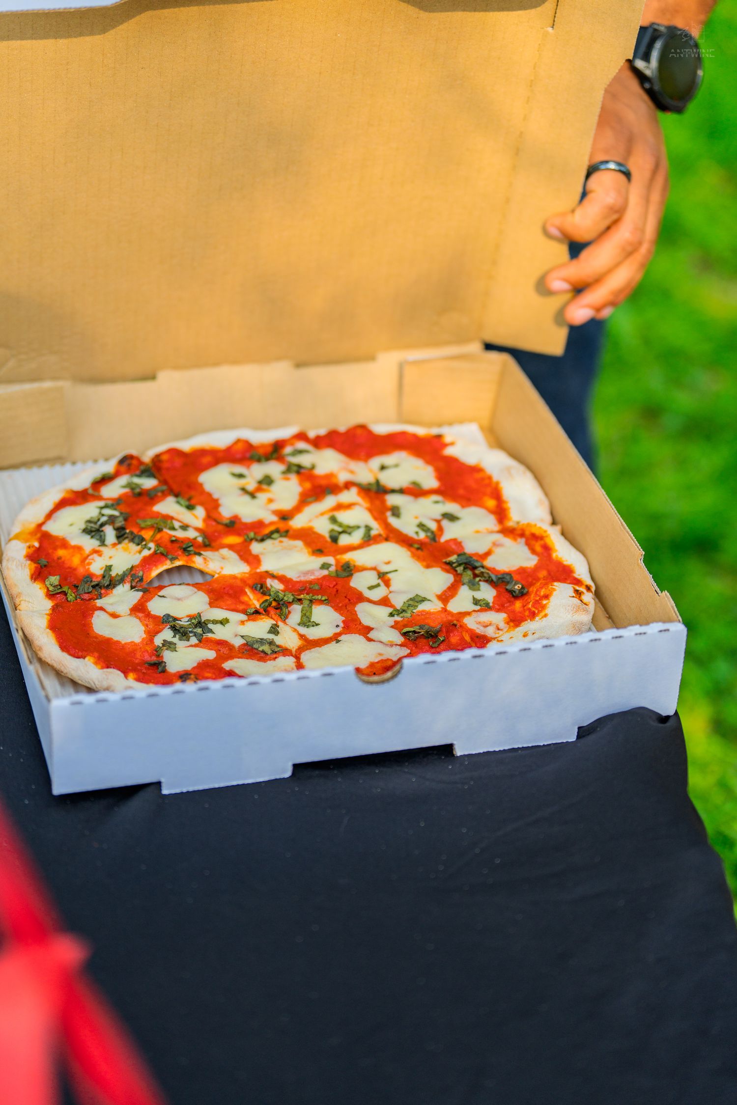 Fresh Margherita pizza in an open delivery box held by a person wearing a watch outdoors.