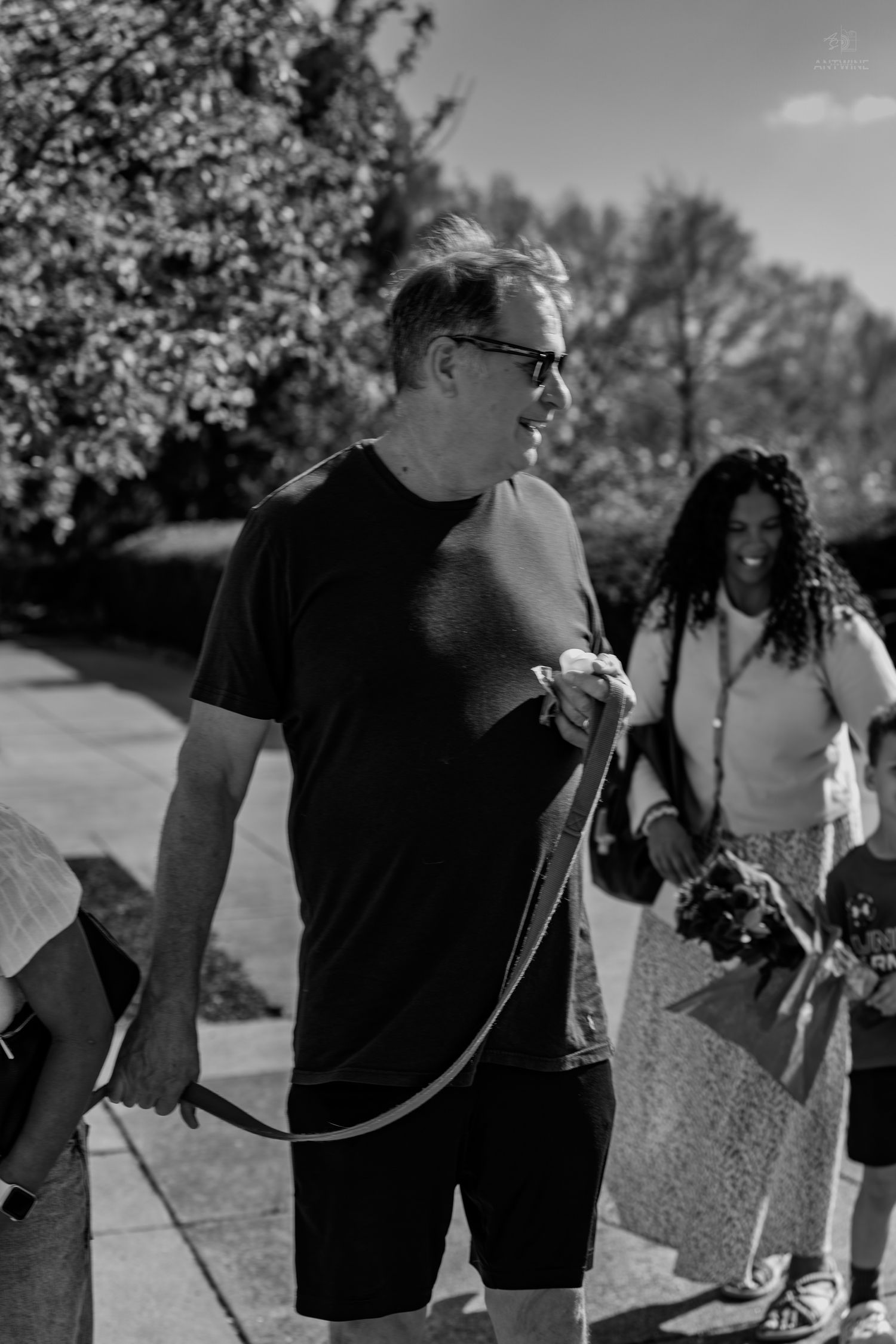 Man in black outfit walks with a dog leash outdoors while a smiling woman with flowers follows behind.