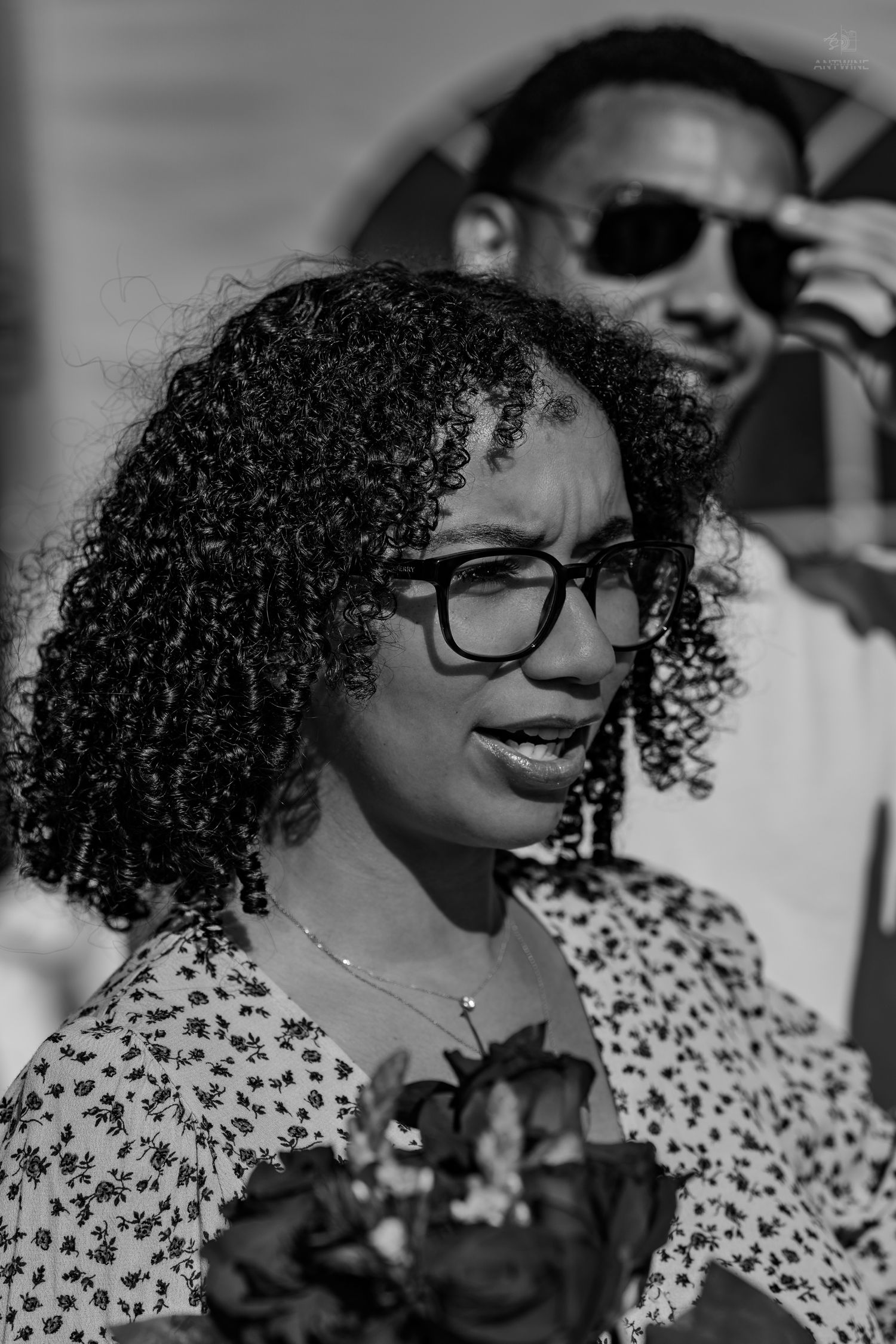 Black and white portrait of a curly-haired woman with glasses holding flowers, man with sunglasses in background.