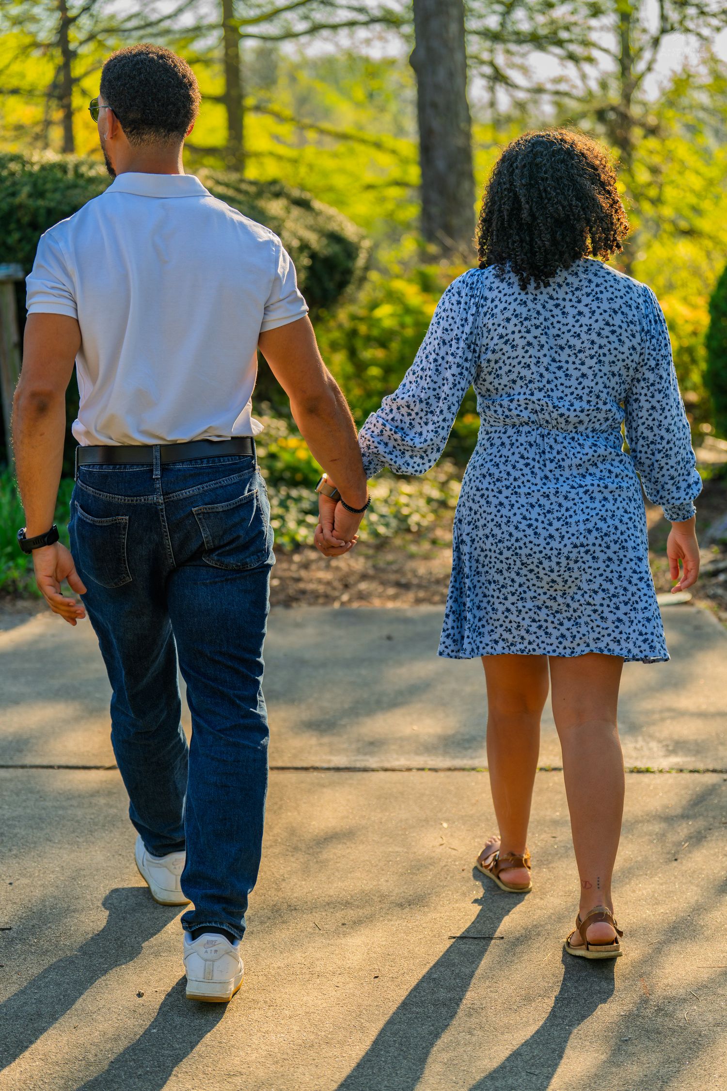 Couple holding hands walking along a sunny garden path, woman in blue floral dress and man in white polo.