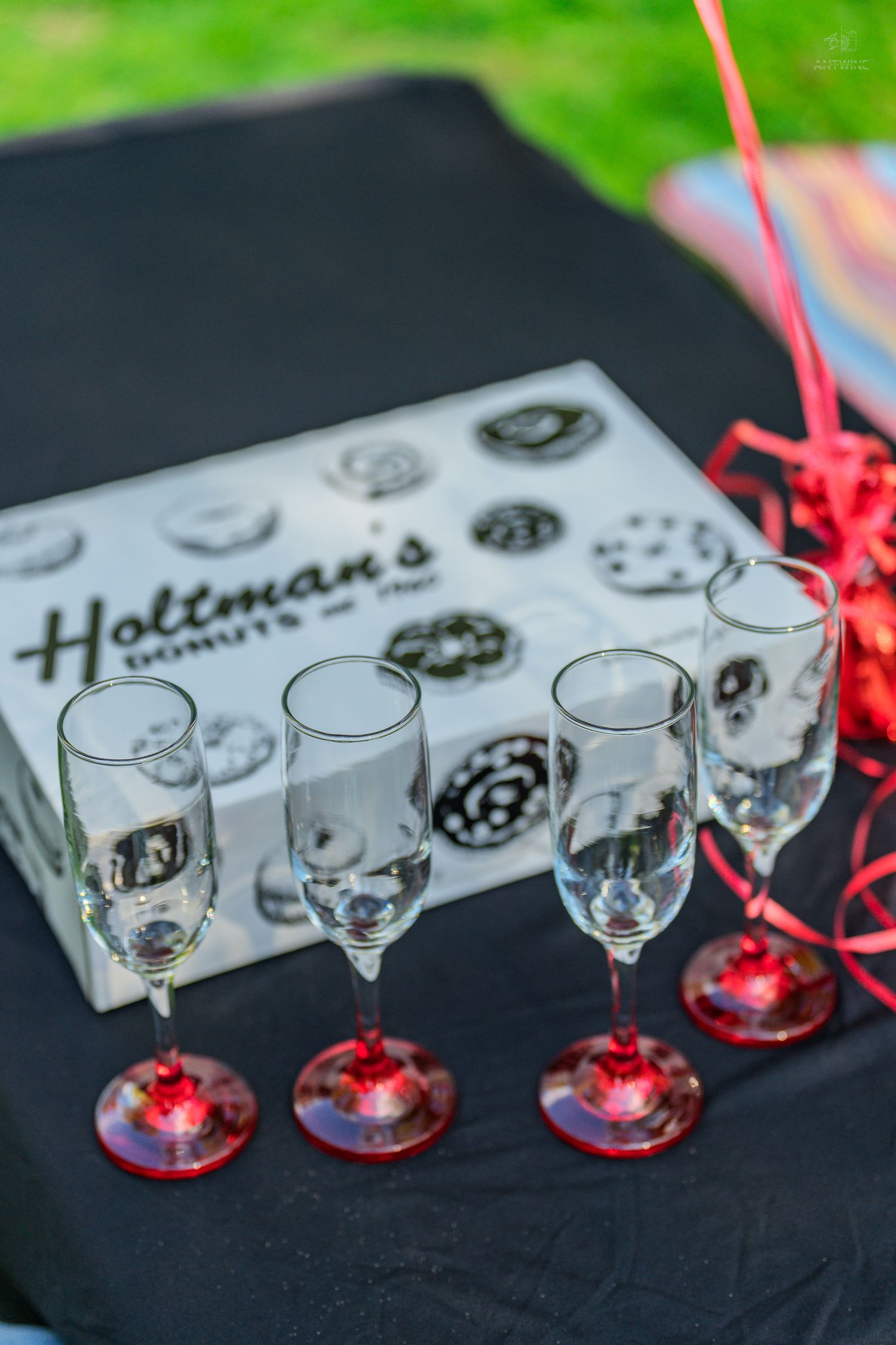 Four champagne flutes with red bases arranged in front of a Holtman's Donuts box on a black tablecloth.