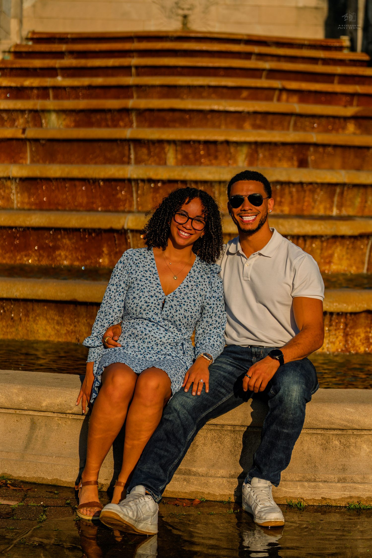 A happy couple sits on sunlit stone steps, the woman in a blue floral dress and man in a white polo shirt.