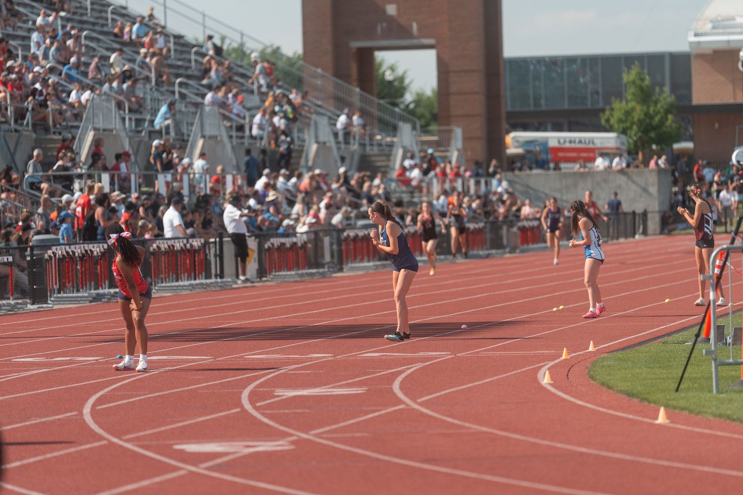 Female athletes compete in a track race at a packed stadium during a high school track and field meet.