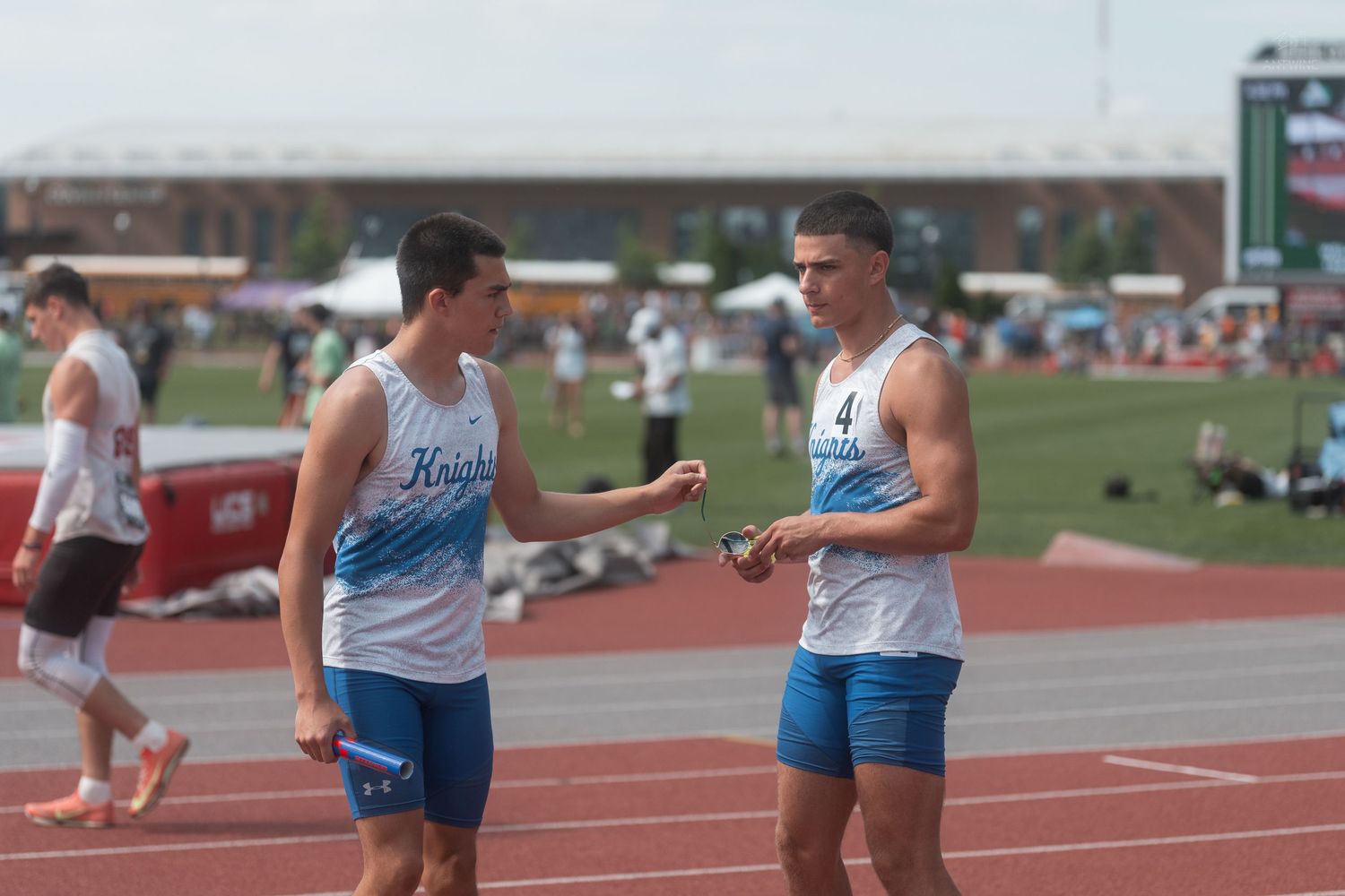 Two male Knights track athletes in blue uniforms converse on track at outdoor field competition.