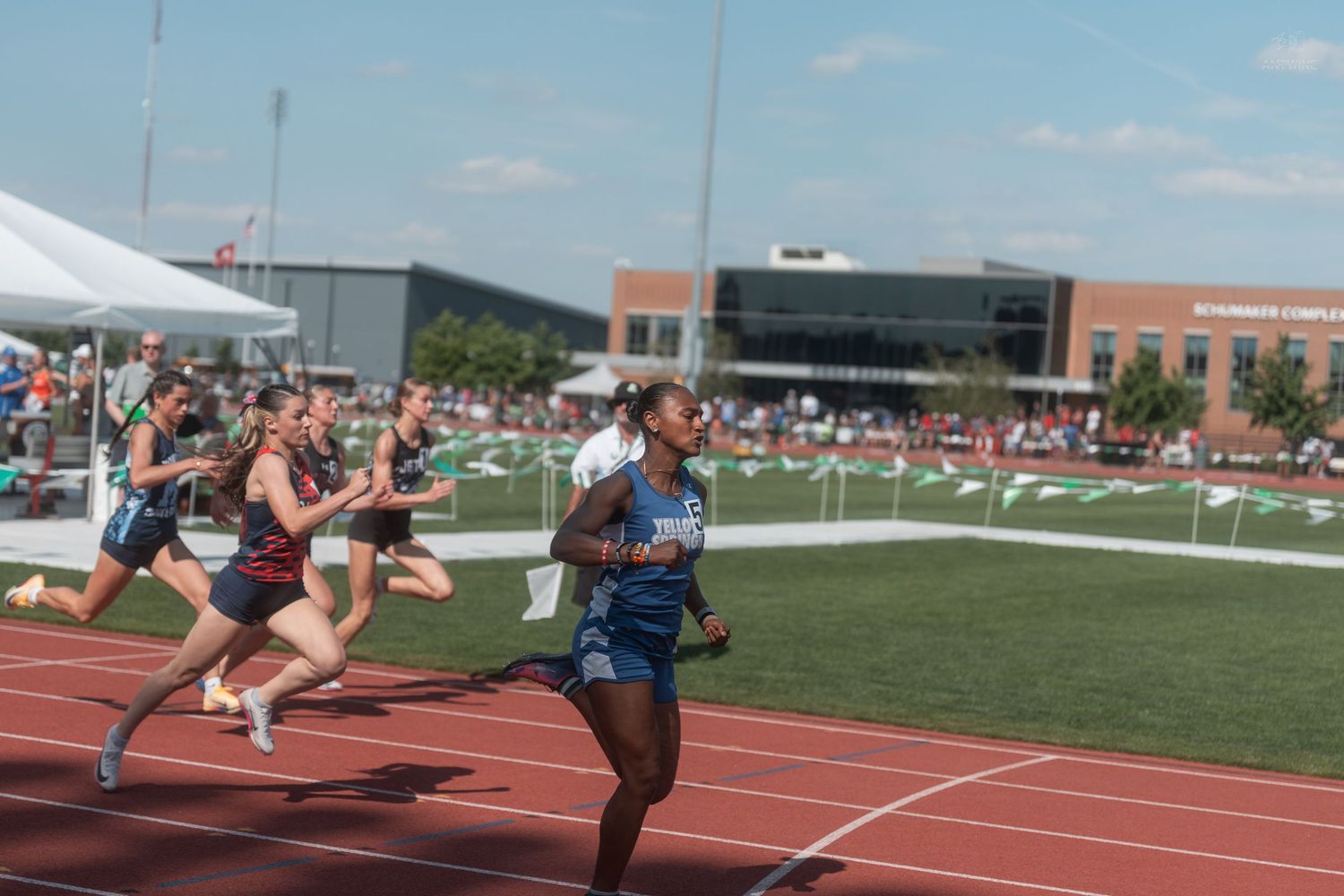A female sprinter in blue leads competitors on a red track at an outdoor high school track and field meet.