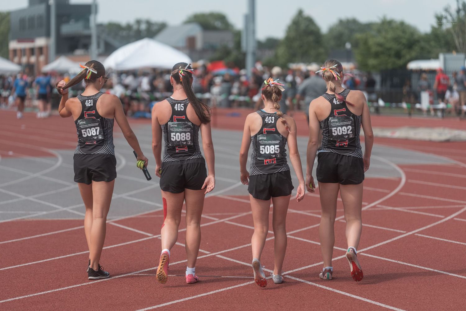 Four female track athletes walk together on a red track wearing matching black uniforms and race numbers.
