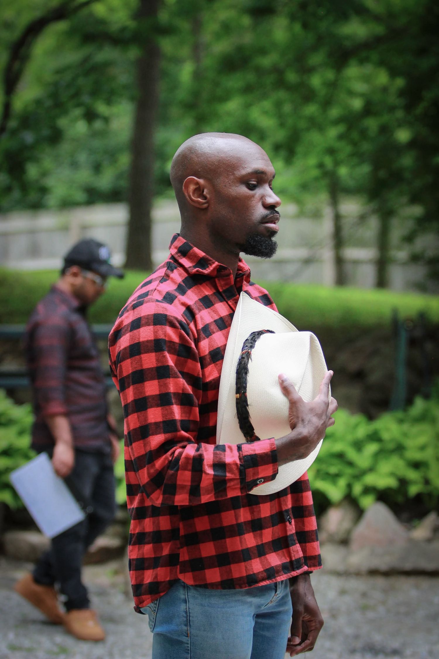 Bald man in red plaid shirt holding a white hat outdoors in a park.