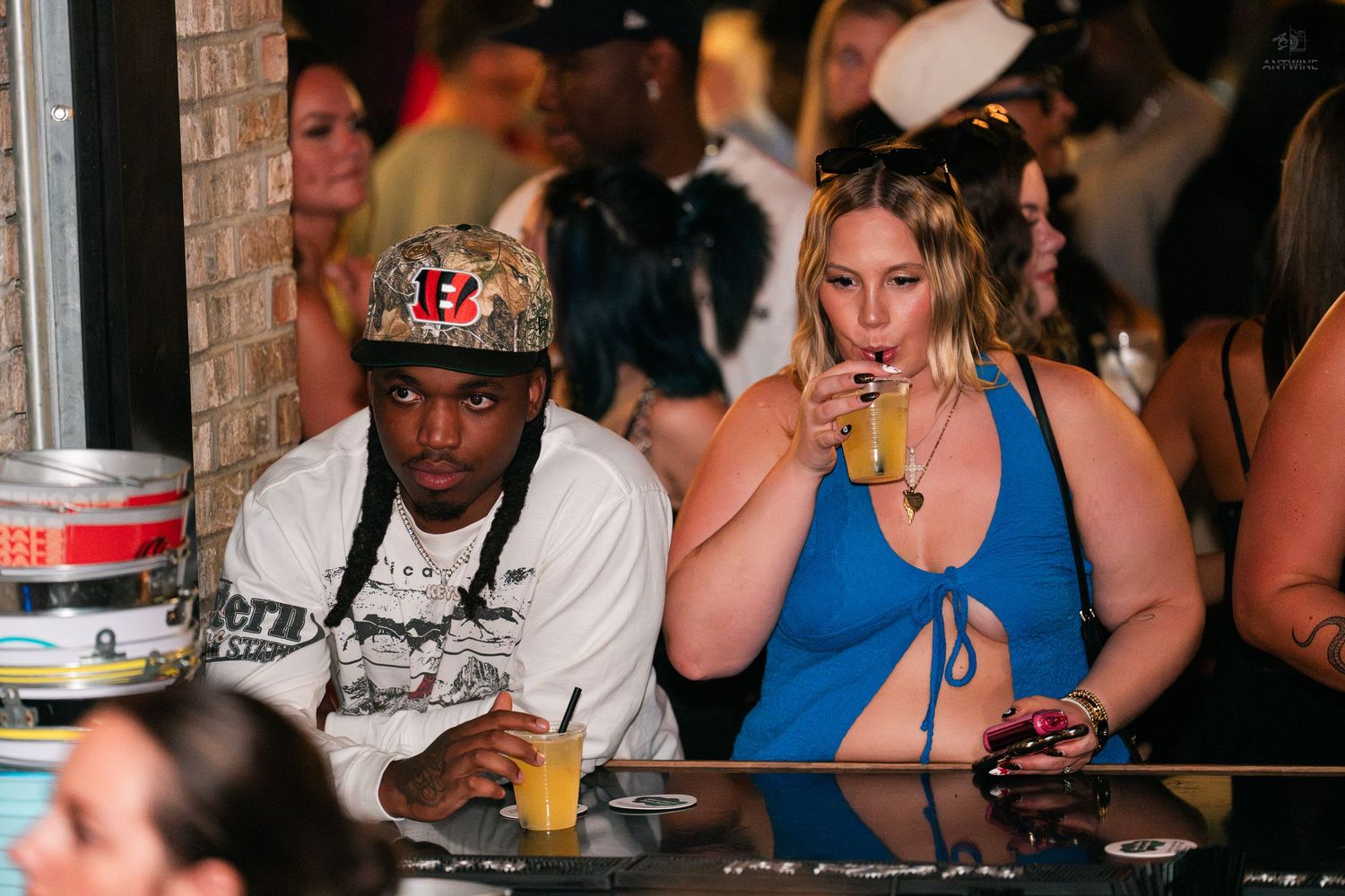 Man in Bengals cap and woman in blue dress enjoying drinks at nightclub bar.