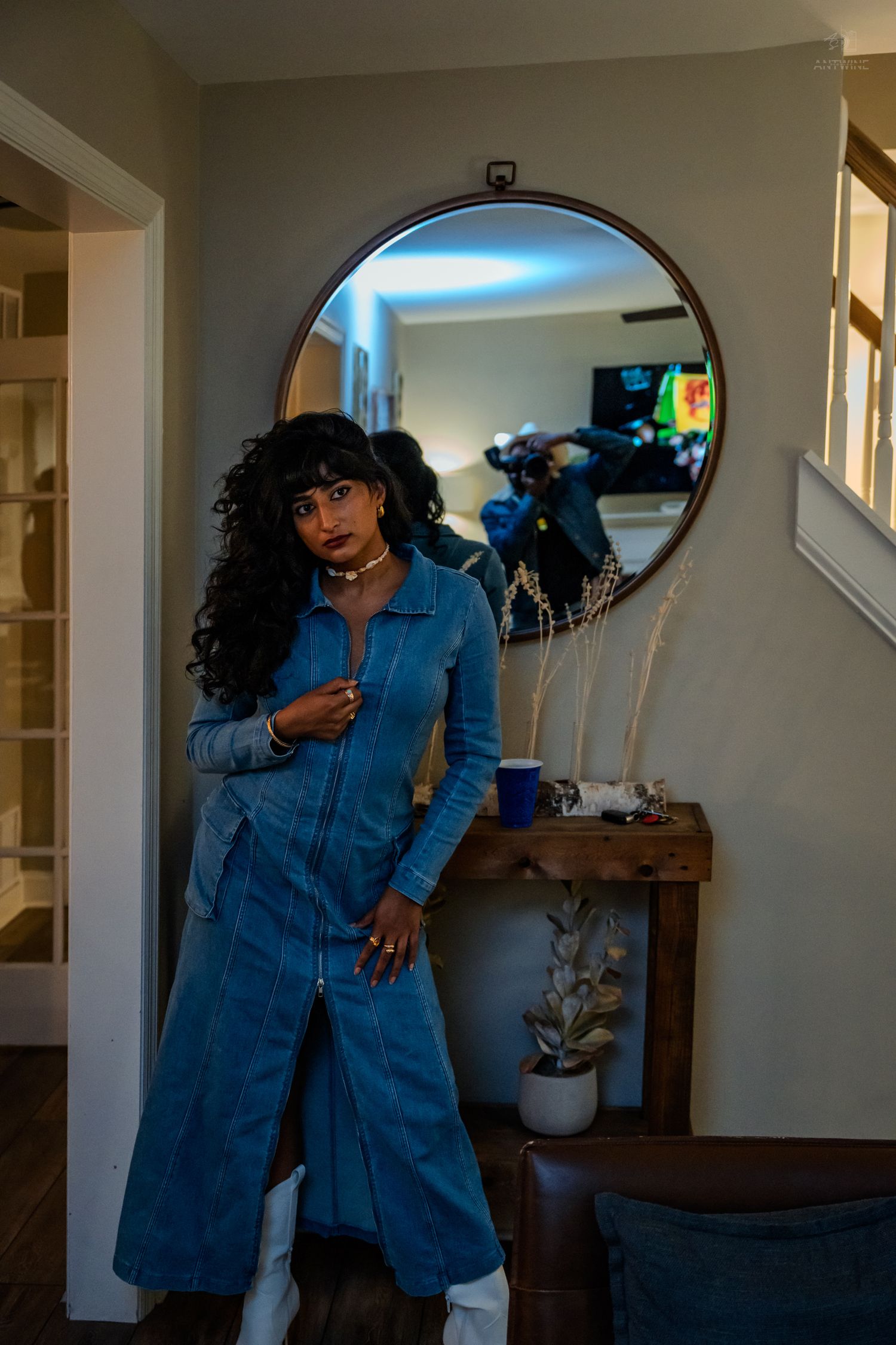 Woman in blue denim dress with curly hair posing in front of round mirror in home hallway.