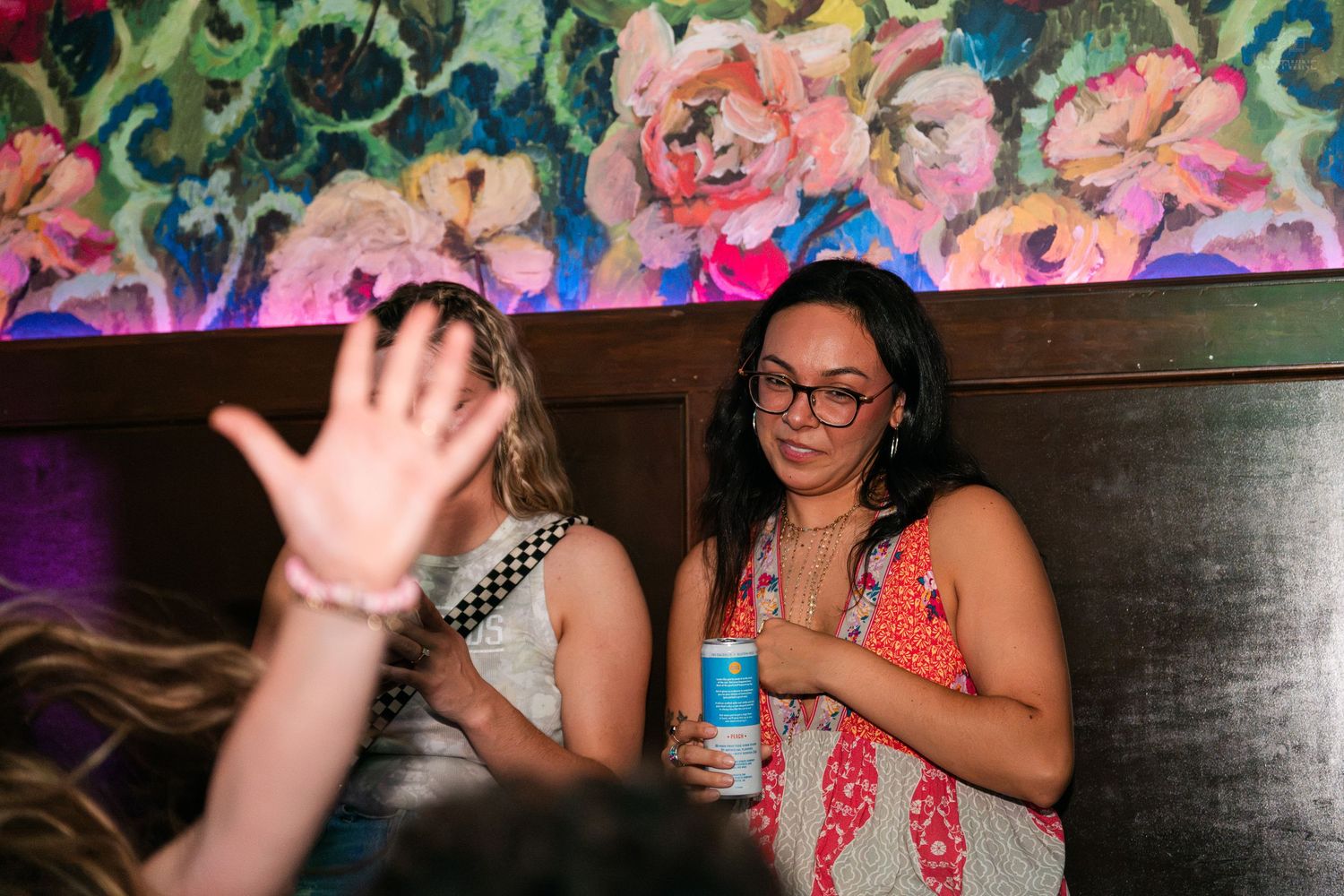 Woman in floral dress holding a canned drink laughing at a lively bar event.