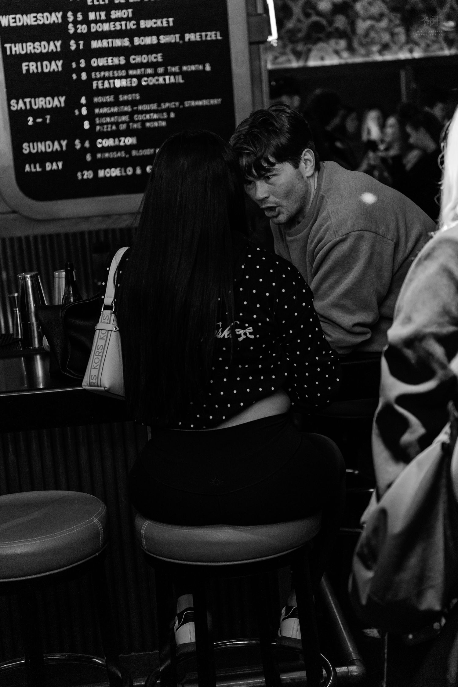 Man and woman having an intimate conversation at a bar with a weekly specials menu board behind them.