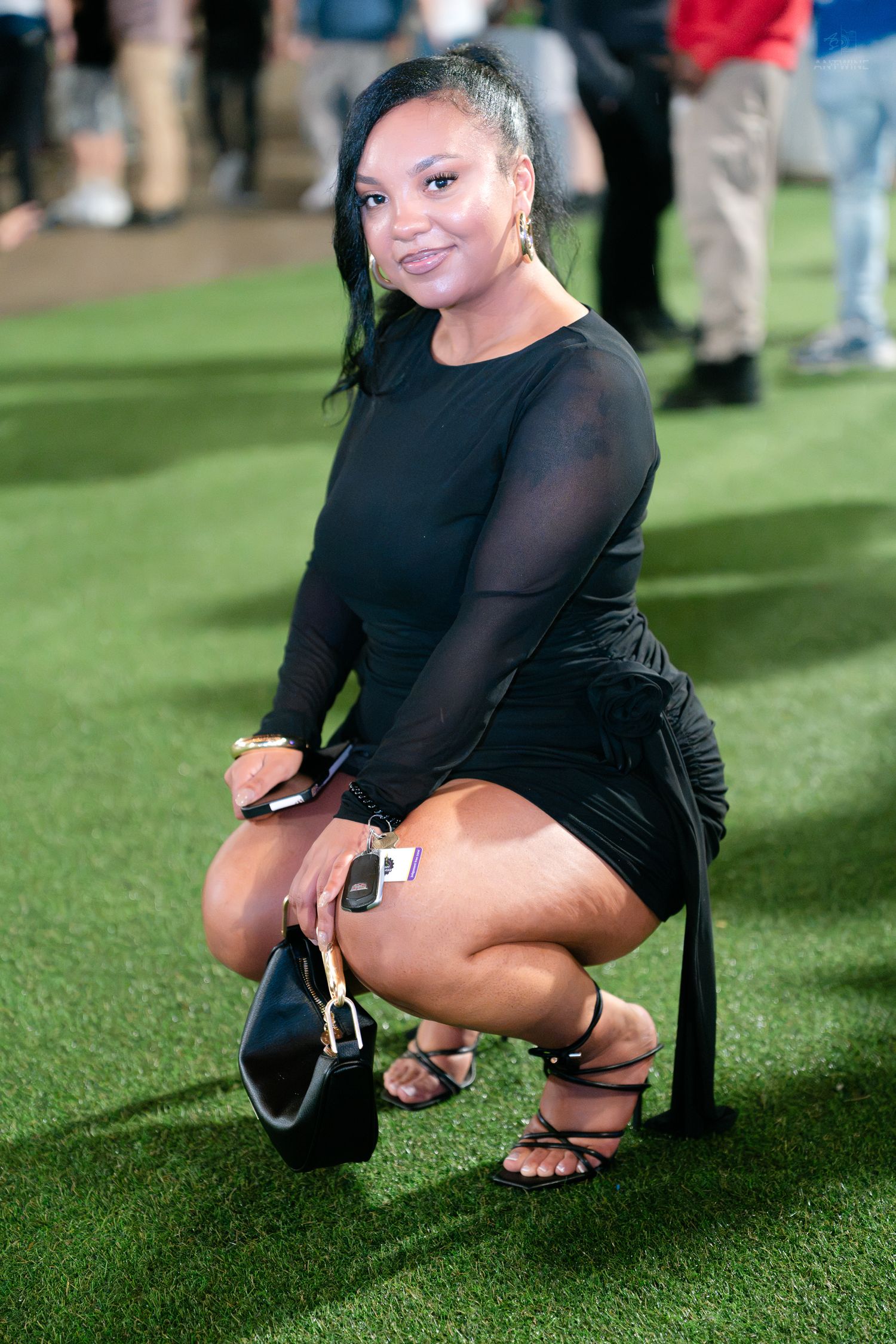 A woman in a black mesh mini dress crouches on artificial turf holding a black handbag.