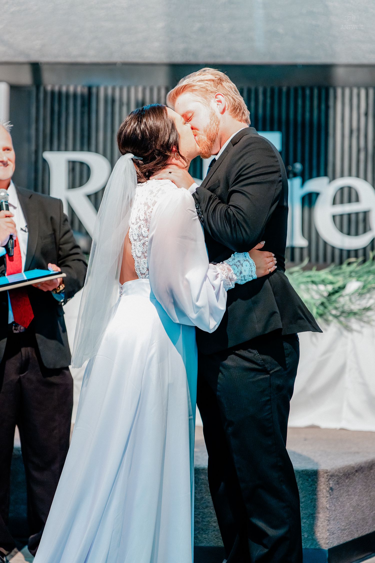 Bride and groom share their first kiss at outdoor wedding ceremony, officiant standing nearby.