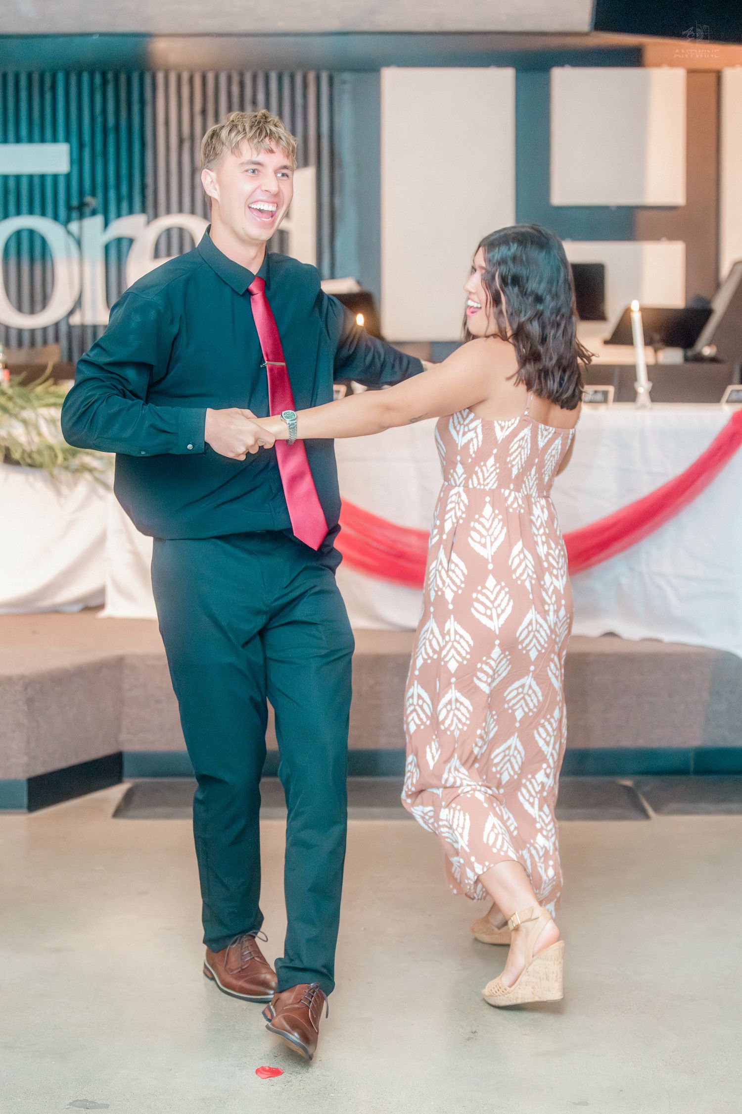 Joyful couple dancing together at wedding reception, man in black shirt with red tie, woman in floral dress.
