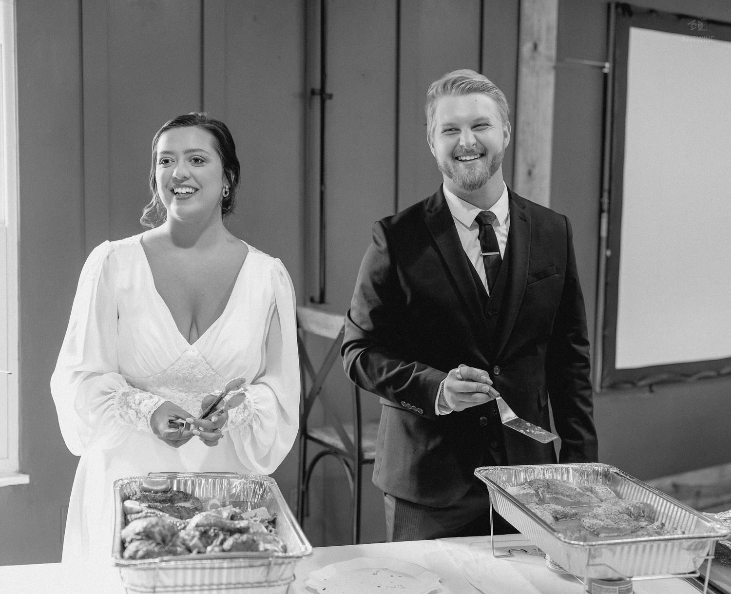Smiling bride and groom serve food at wedding reception buffet table in black and white photo.