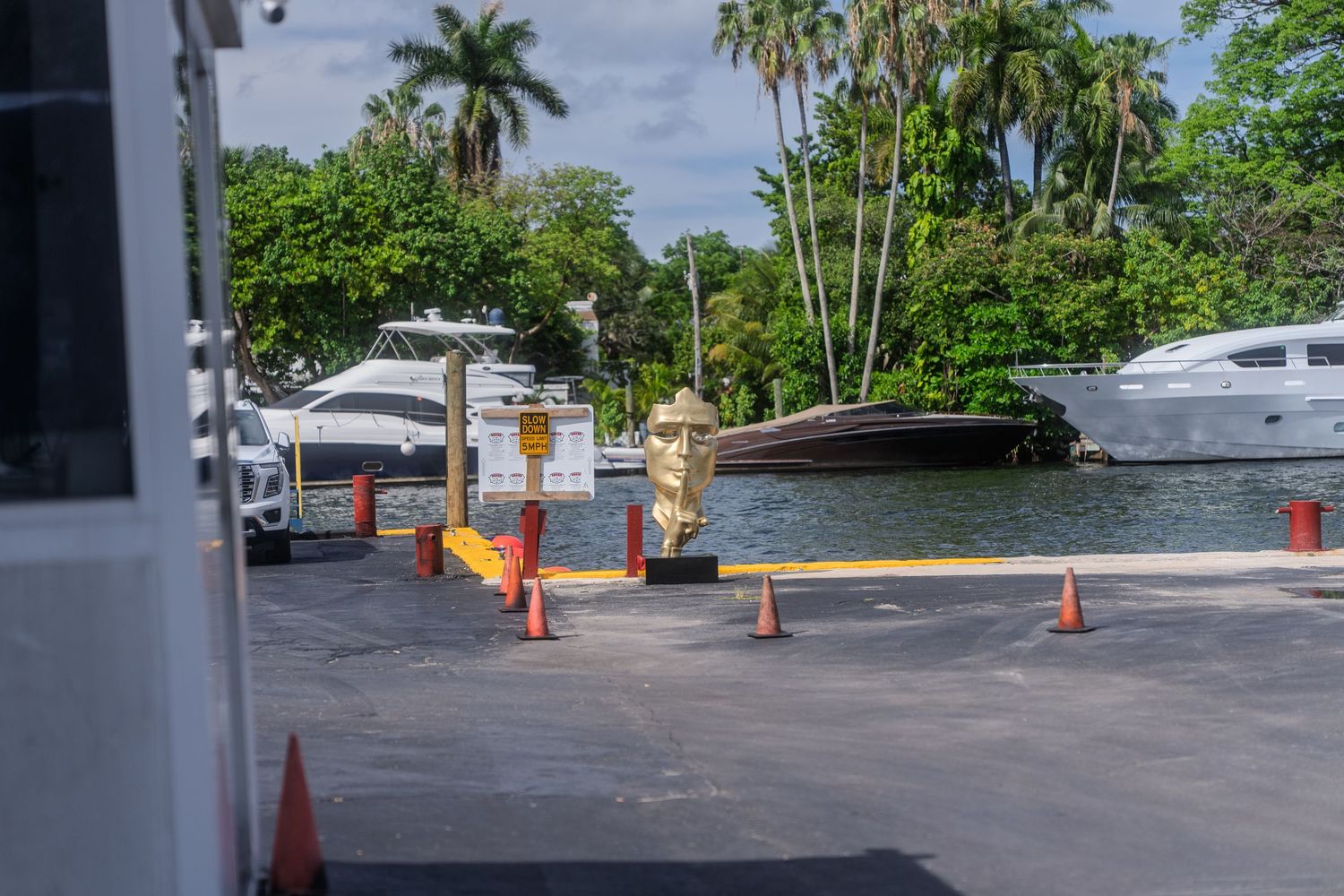 A marina dock with orange traffic cones and luxury yachts surrounded by tropical palm trees.