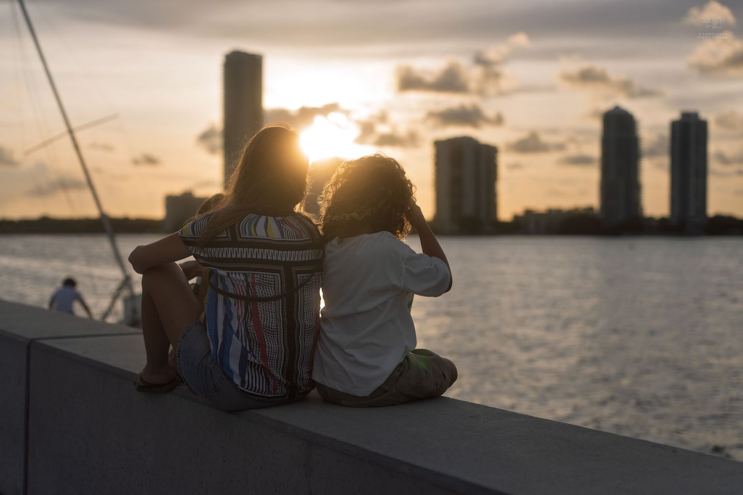 Two friends sitting together on a waterfront ledge watching a beautiful golden sunset over the city skyline.
