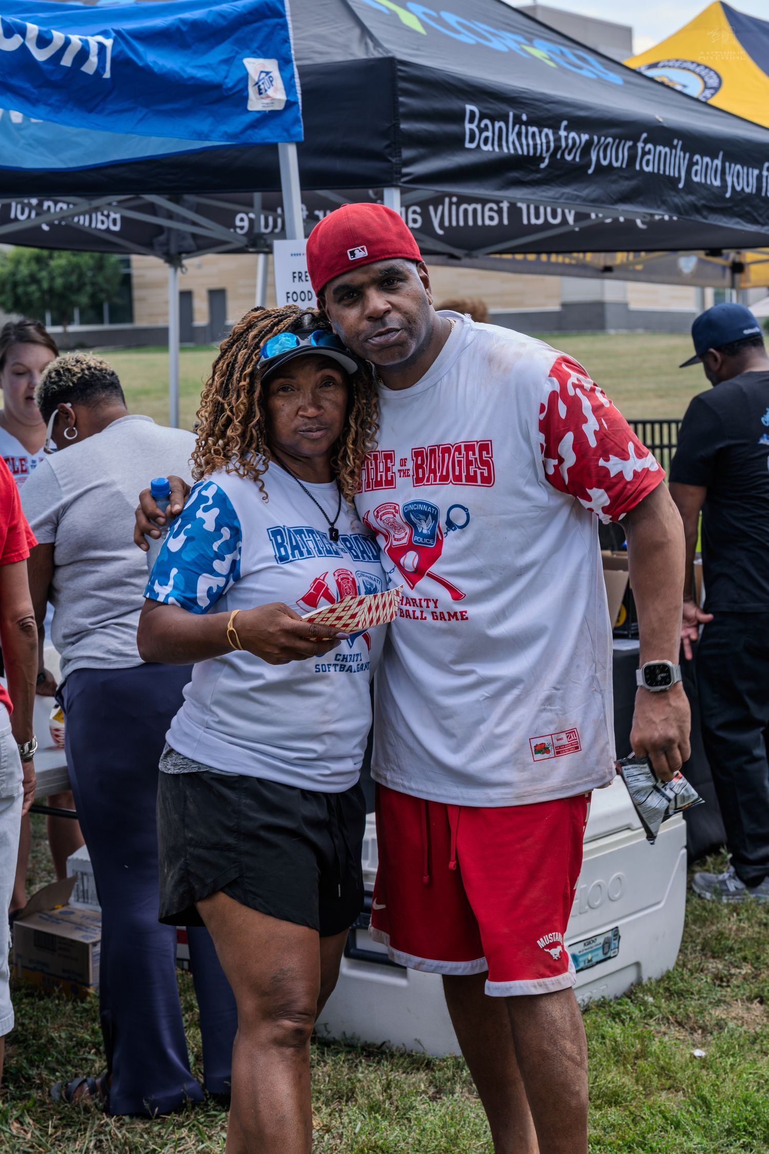 Couple posing together at outdoor charity softball game event under banking sponsor tent on sunny day.
