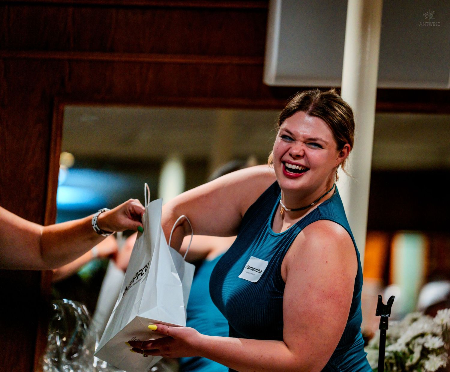 A smiling woman in a teal dress receives a gift bag at a formal event, wearing a name tag.