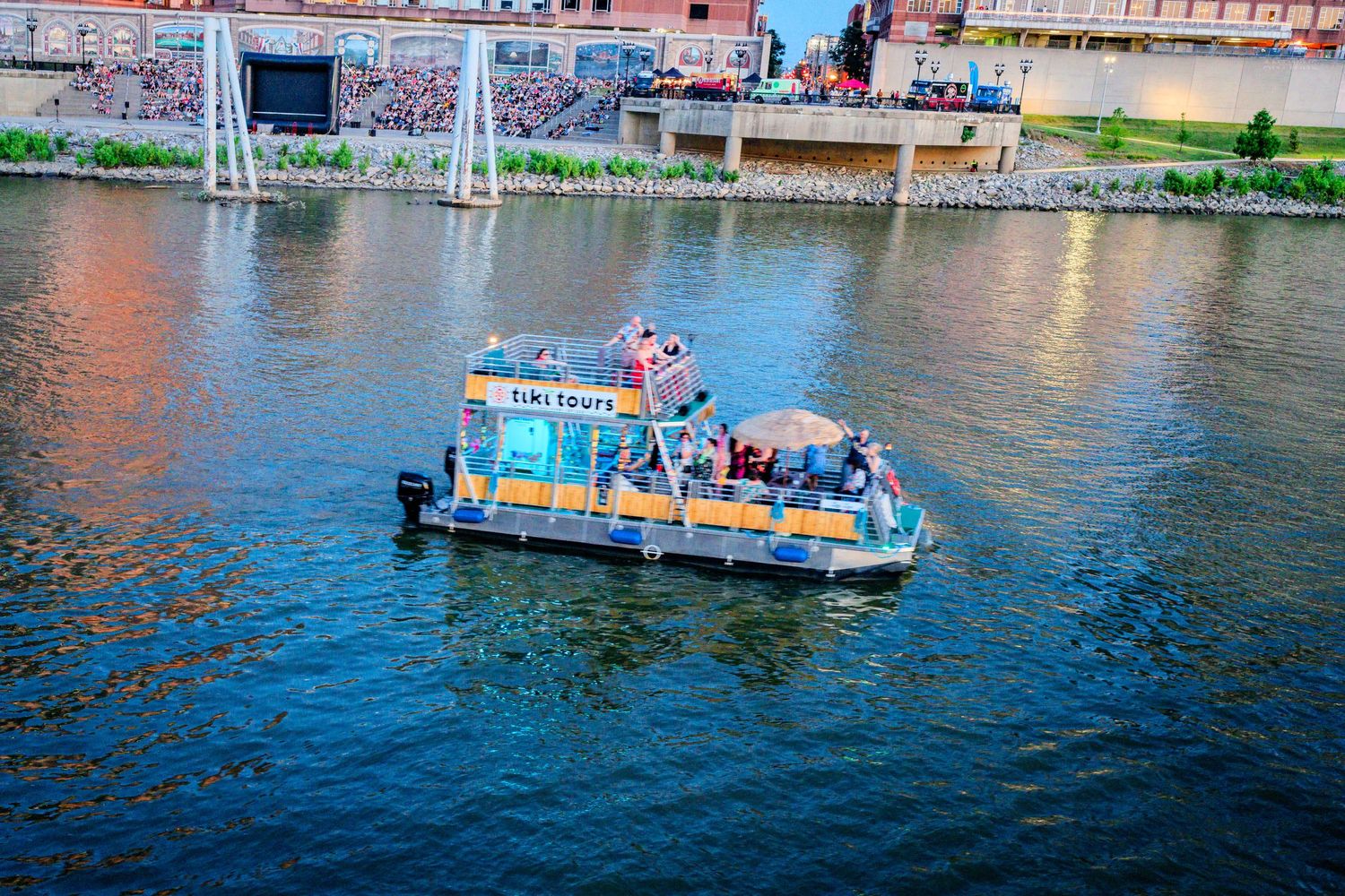 Tiki Tours pontoon boat carrying passengers along a city river at sunset.