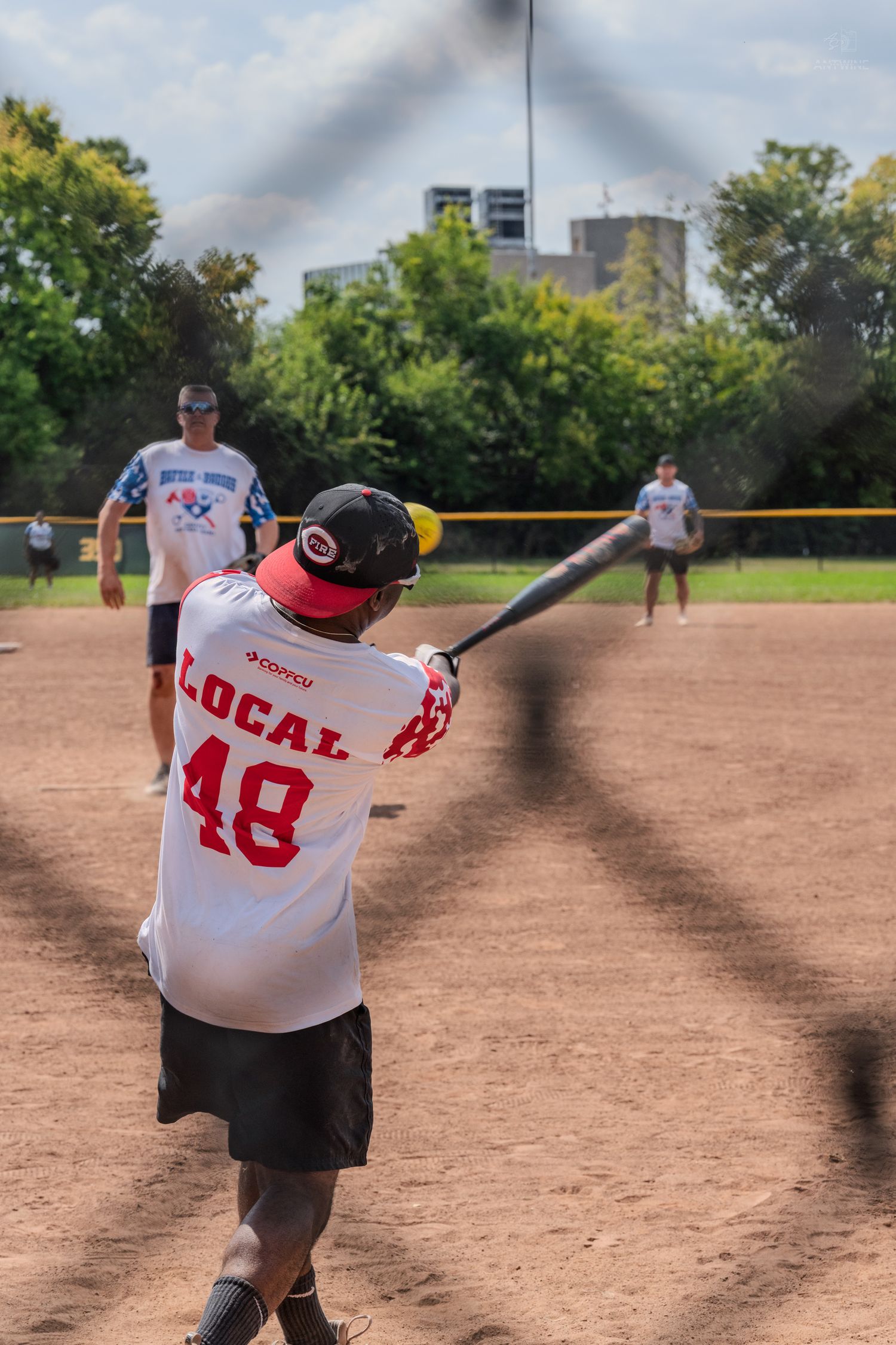 Player in Local 48 jersey hitting a softball during an outdoor daytime game viewed through chain-link fence.