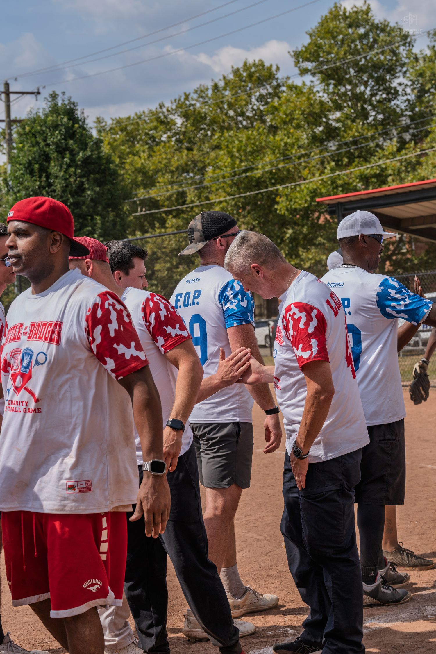 Group of men in matching white and red softball jerseys lining up on a dirt baseball field during a charity game.