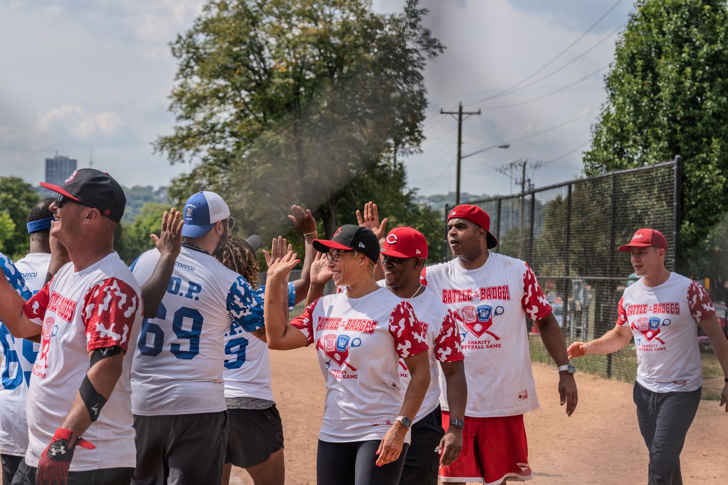Team members in matching softball jerseys celebrate on a dusty field during a charity softball game on a sunny day.