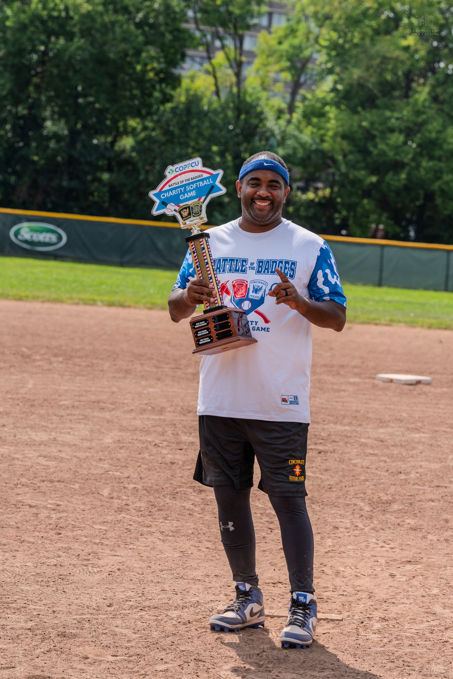 Smiling man proudly holds a Battle of Badges Charity Softball Game trophy on the field.
