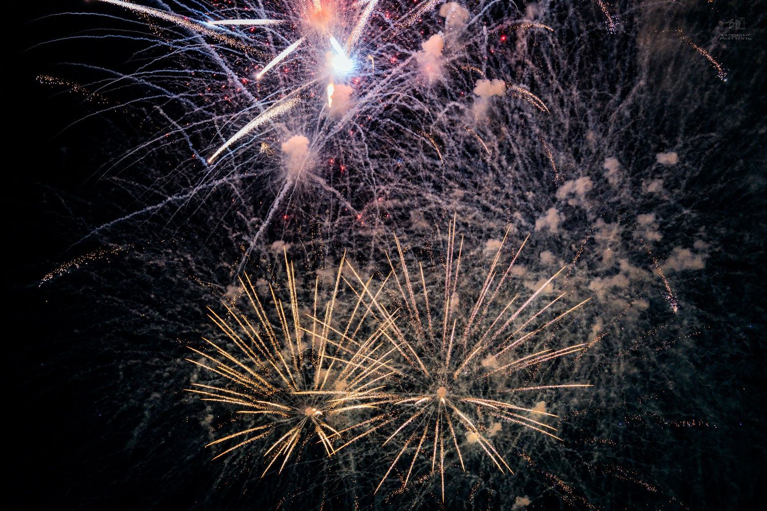 Brilliant gold and blue fireworks exploding against a dark night sky with smoke trails.