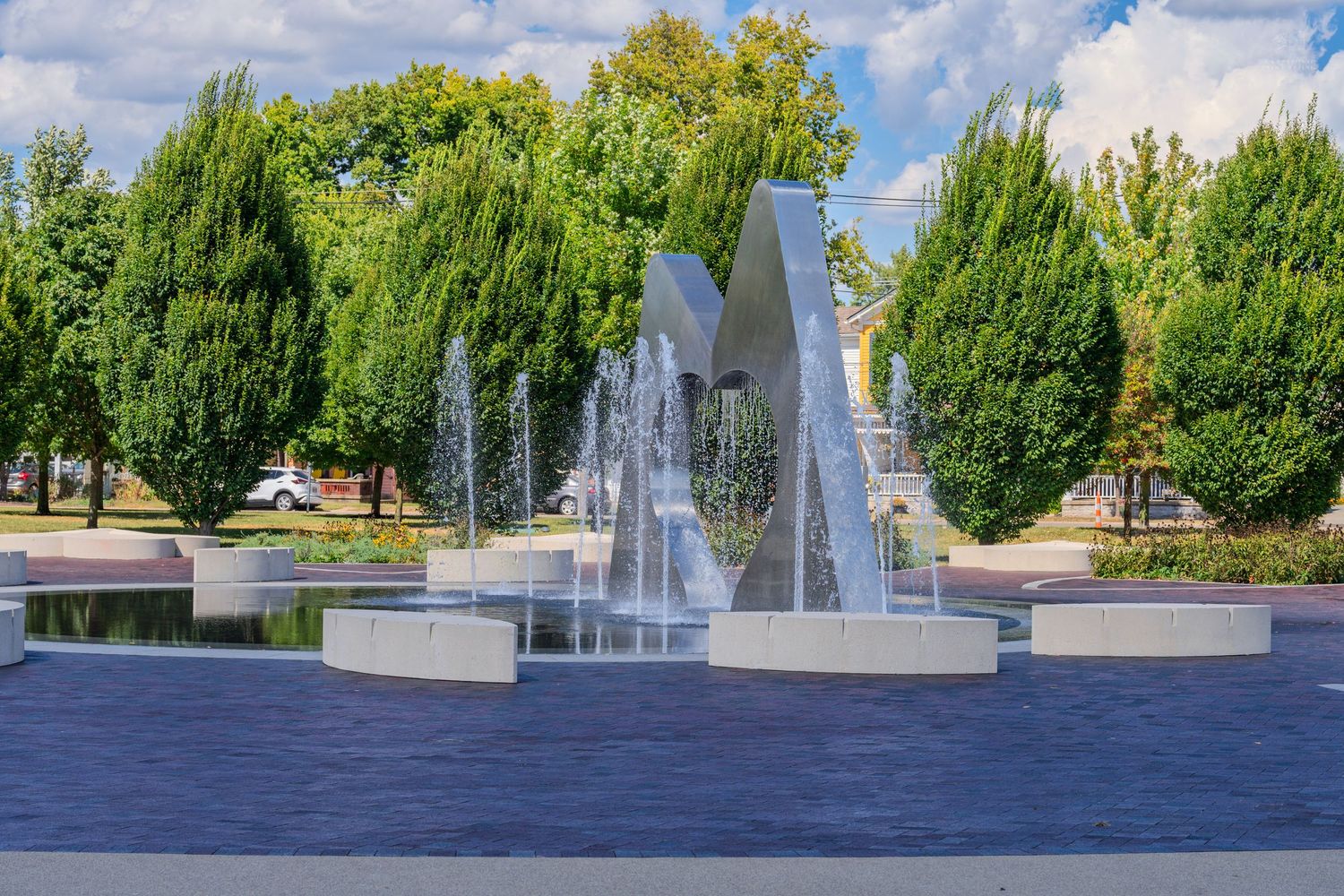 Outdoor fountain with metal sculpture surrounded by water jets, lush green trees, and a brick plaza under a clear blue sky.