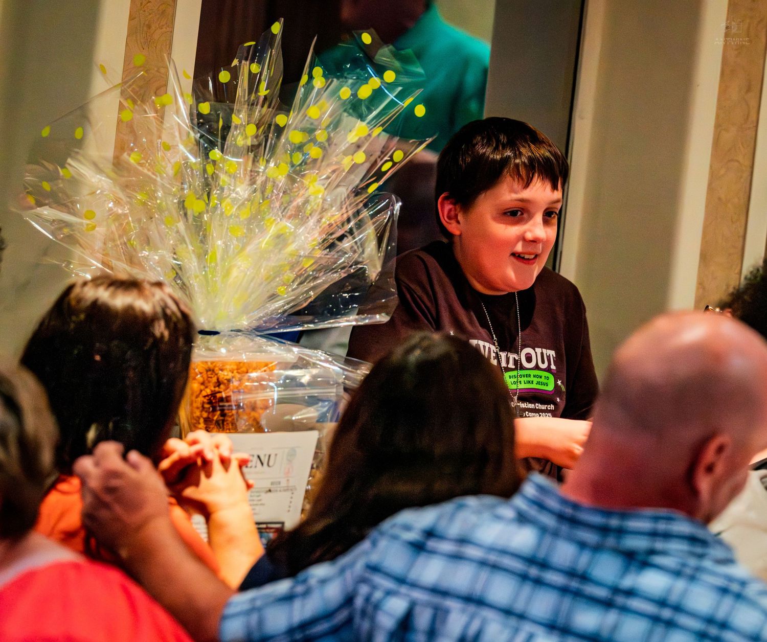 A young boy in a black shirt smiles proudly beside a large wrapped gift basket at an indoor event.