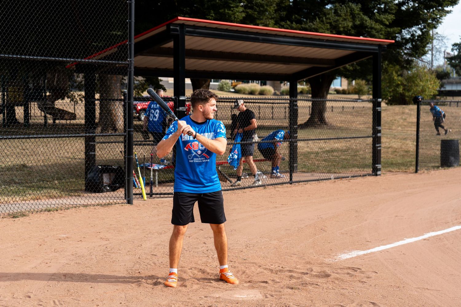 A softball batter in a blue uniform stands ready at home plate on a sunny dirt field.