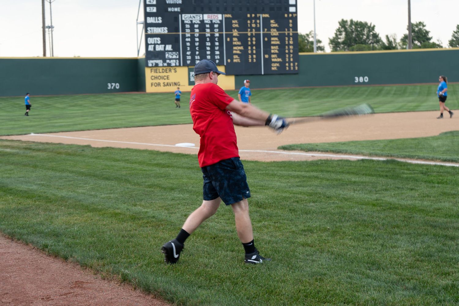 Man in red shirt swinging baseball bat on green field with scoreboard in background.