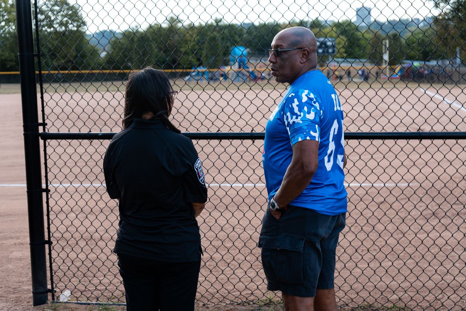Two spectators watch a softball game through a chain-link fence at an outdoor sports field.