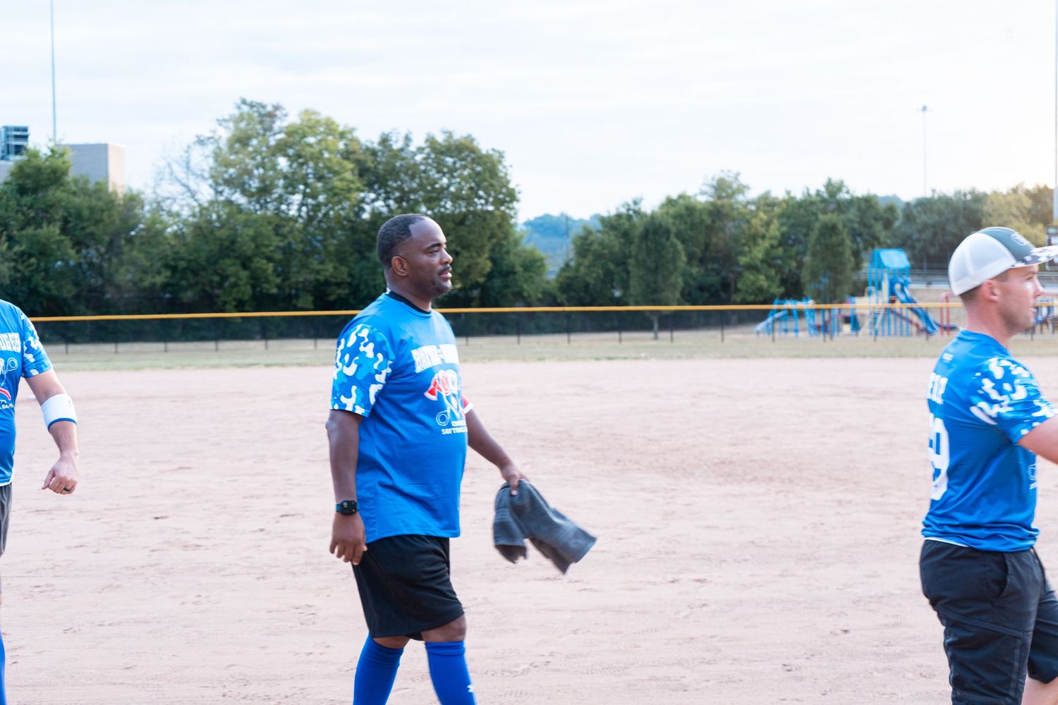 A tall man in a blue jersey holds a mitt on a sandy softball field during an outdoor recreational game.