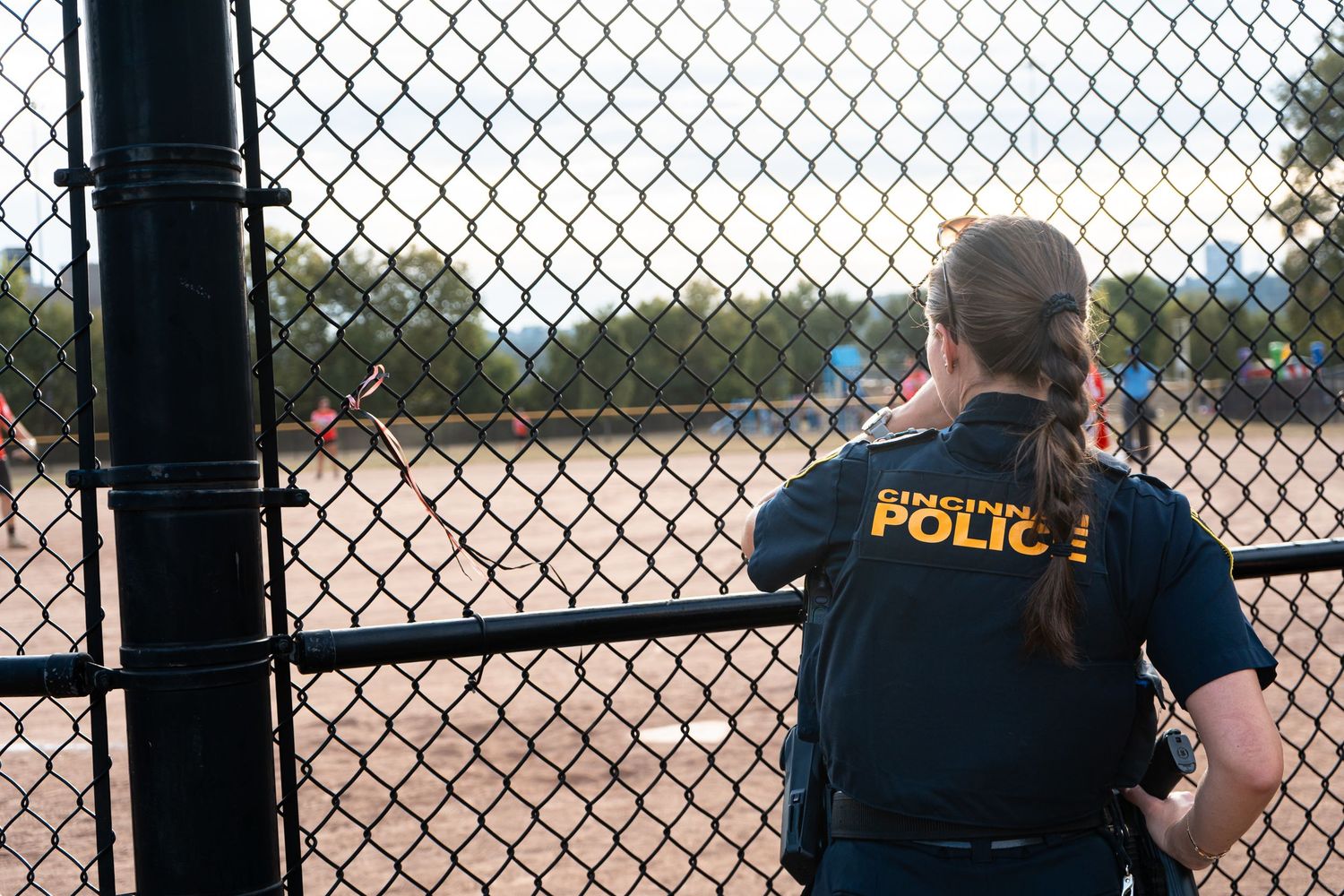 A Georgetown Police officer in uniform watches a softball game through a chain-link fence from behind.