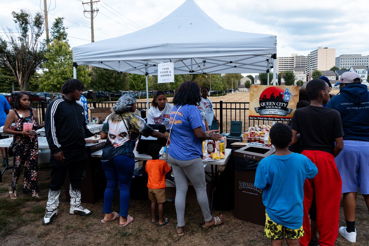Community members gather under a white tent at an outdoor food event in an urban park setting.