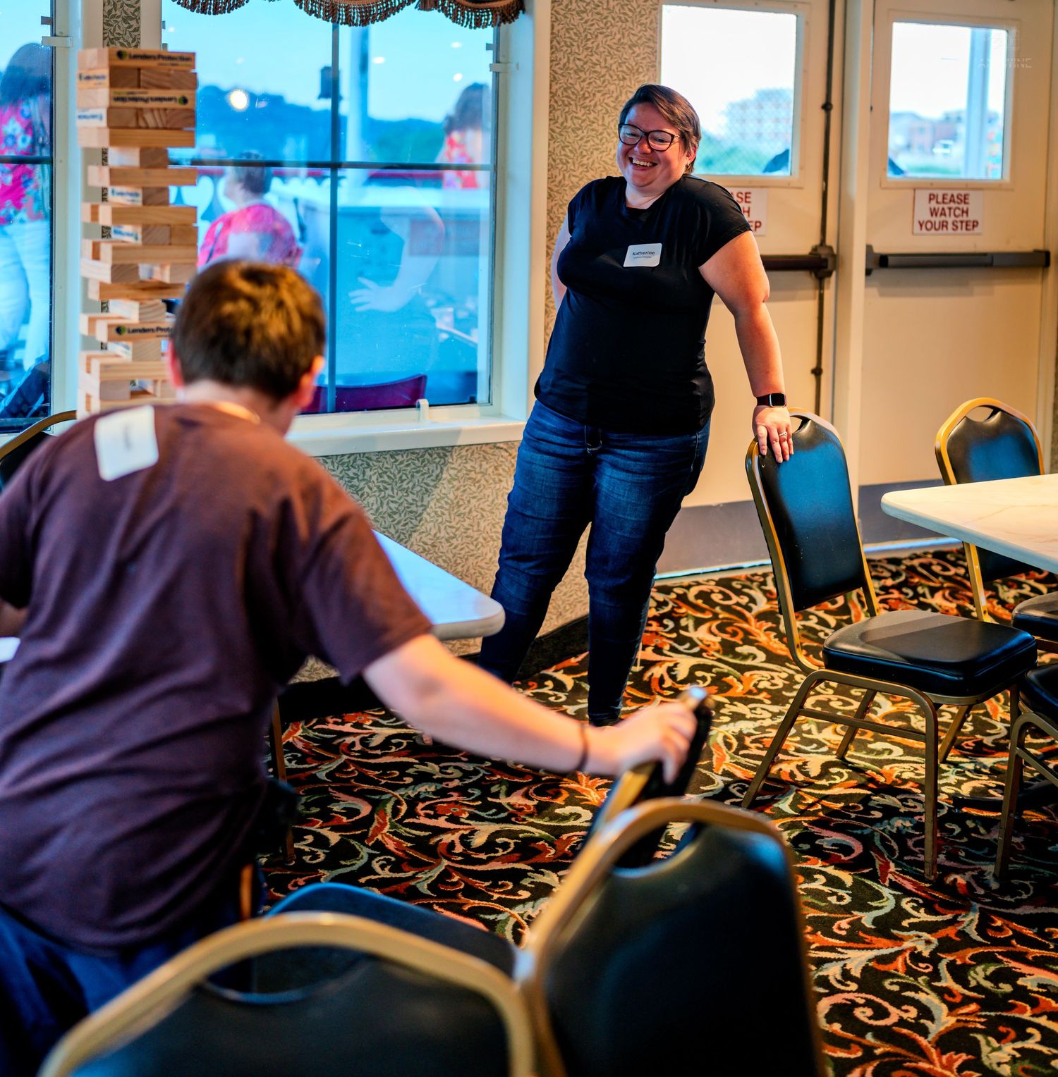 Two people arranging chairs in a banquet hall with patterned carpet during event setup.